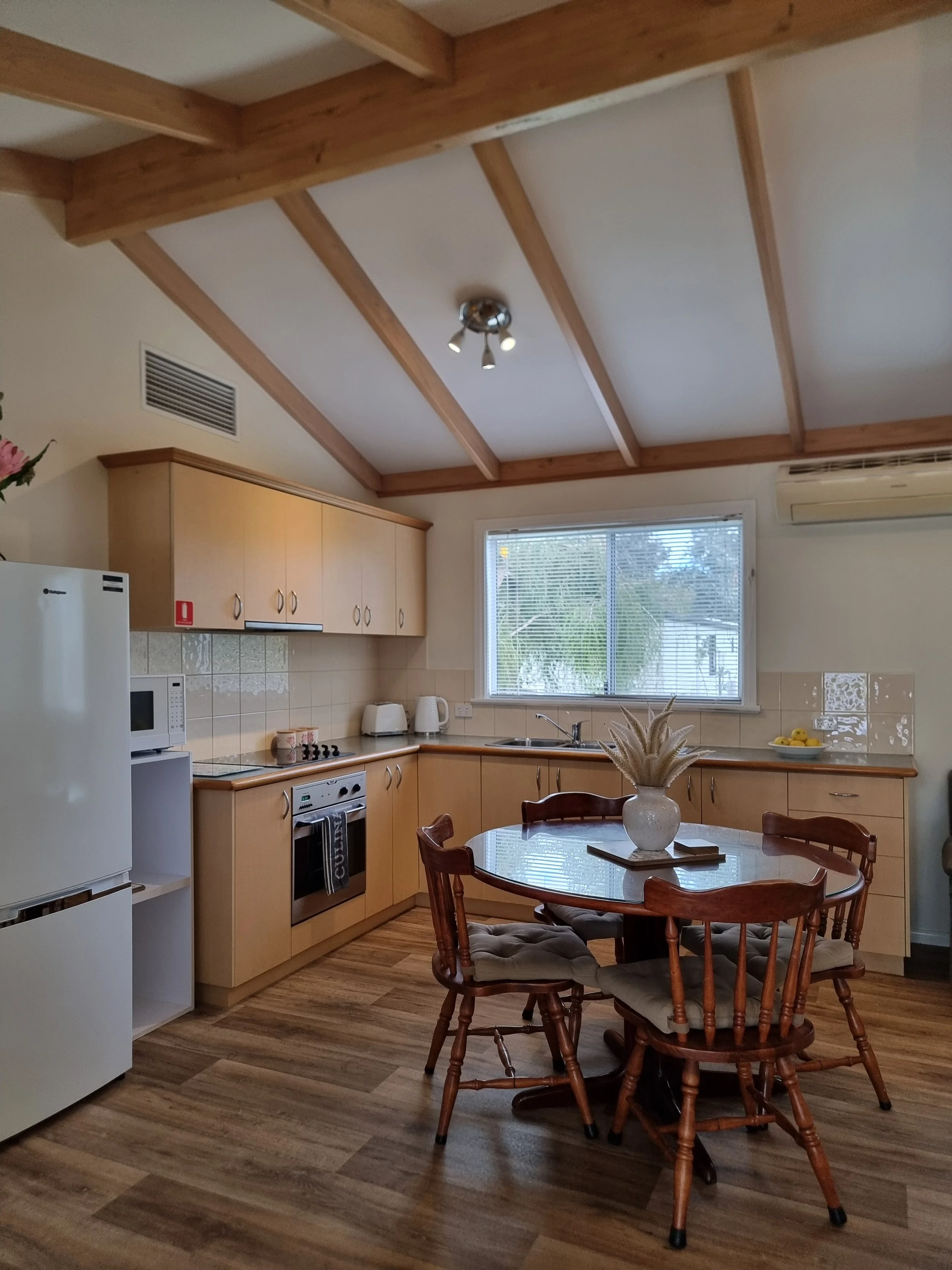 A cozy kitchen with light wooden cabinetry, a round dining table with four chairs, and a window with blinds showing outdoor greenery. The ceiling features exposed wooden beams and a modern ceiling light fixture.
