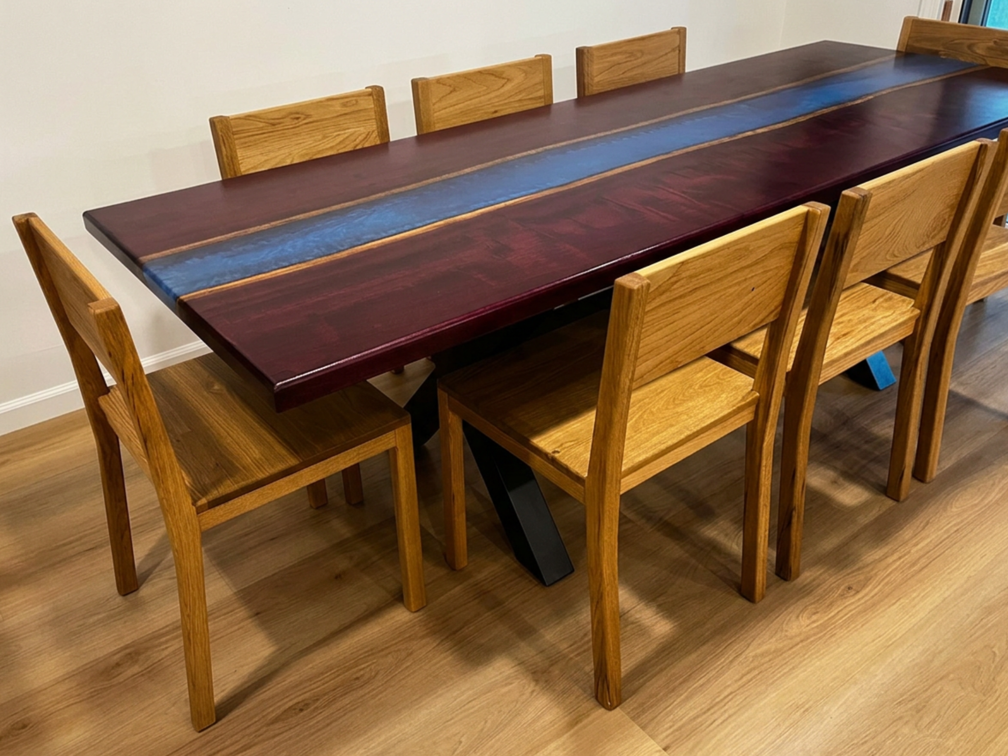 Long wooden dining table with a dark wood top, featuring a central blue resin inlay. Surrounding the table are light-colored wooden chairs with vertical slats on the backrests, set on a wooden floor against a wall.