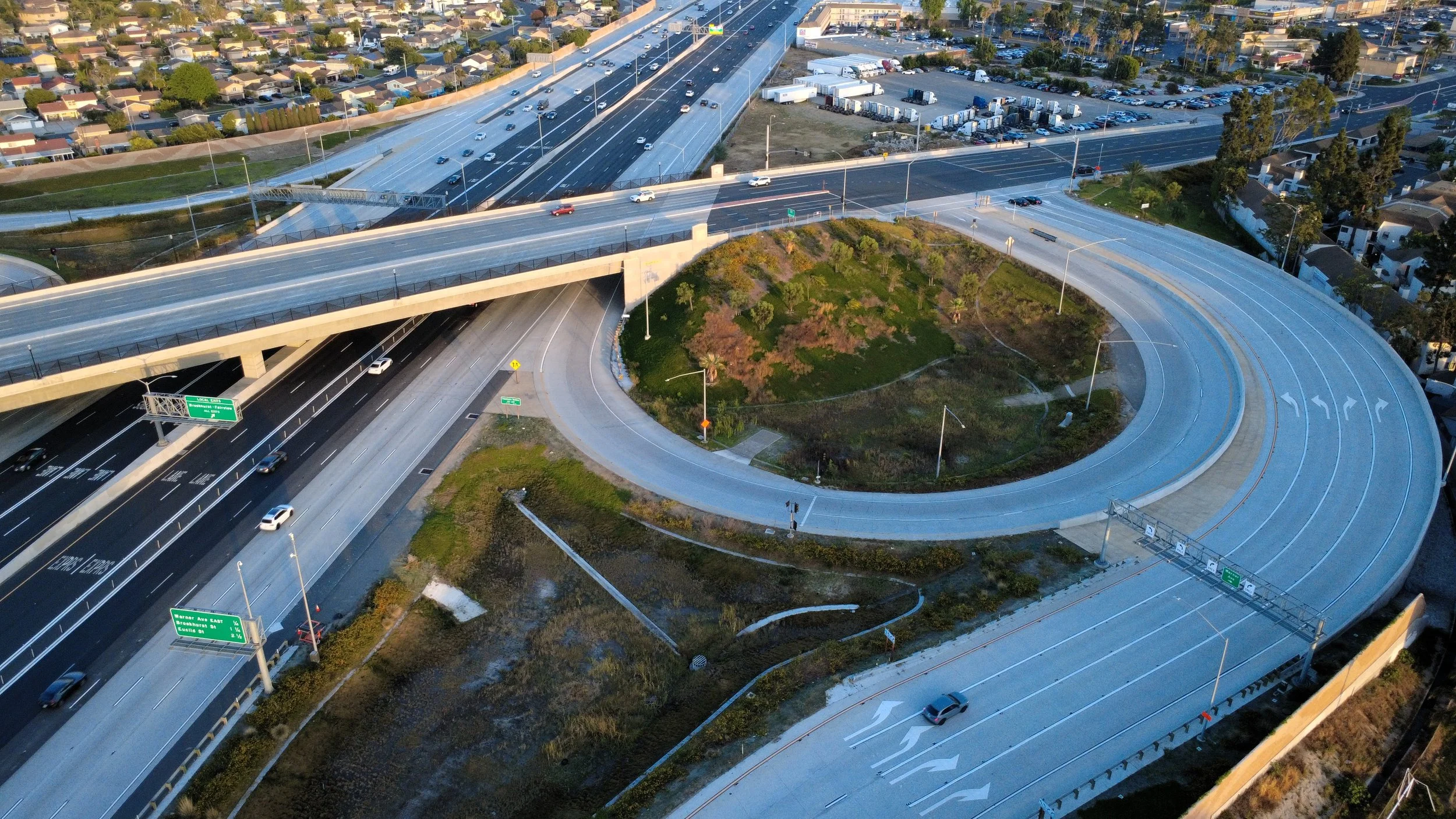 An aerial view of a highway interchange with multiple roads, overpasses, and ramps. Cars are driving on the highways, and there is a grassy area with trees and landscaping in the center of the interchange. Residential houses and parking lots are visible in the background.