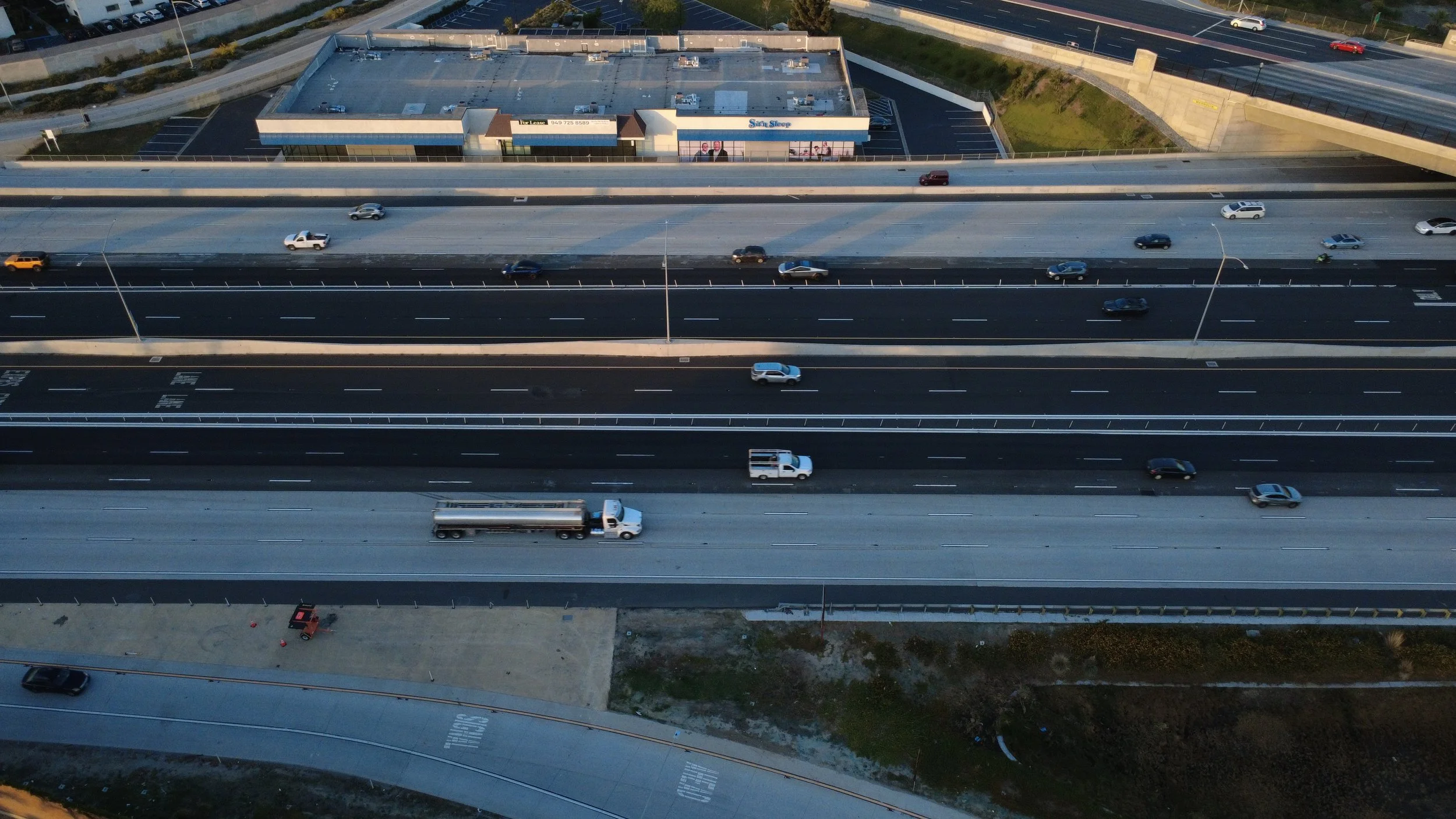An aerial view of a multi-lane highway with several vehicles, including cars and trucks, traveling in both directions. In the background, there's a commercial building with a parking lot and an overpass.