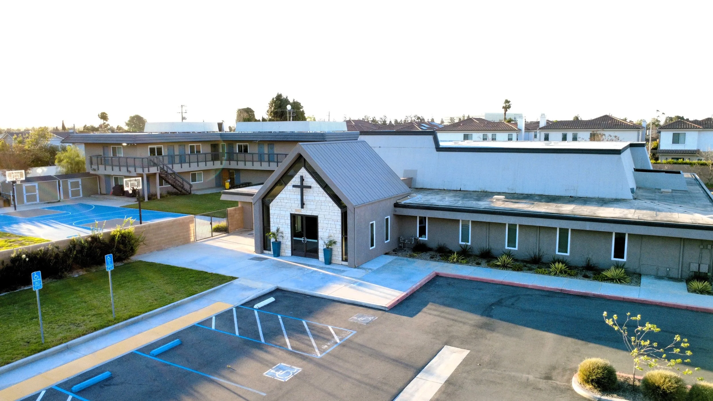 A church or community building with a parking lot and basketball courts. The building has a cross on the front, glass doors, and a modern design with a sloped roof. There are designated handicapped parking spaces and outdoor plants.
