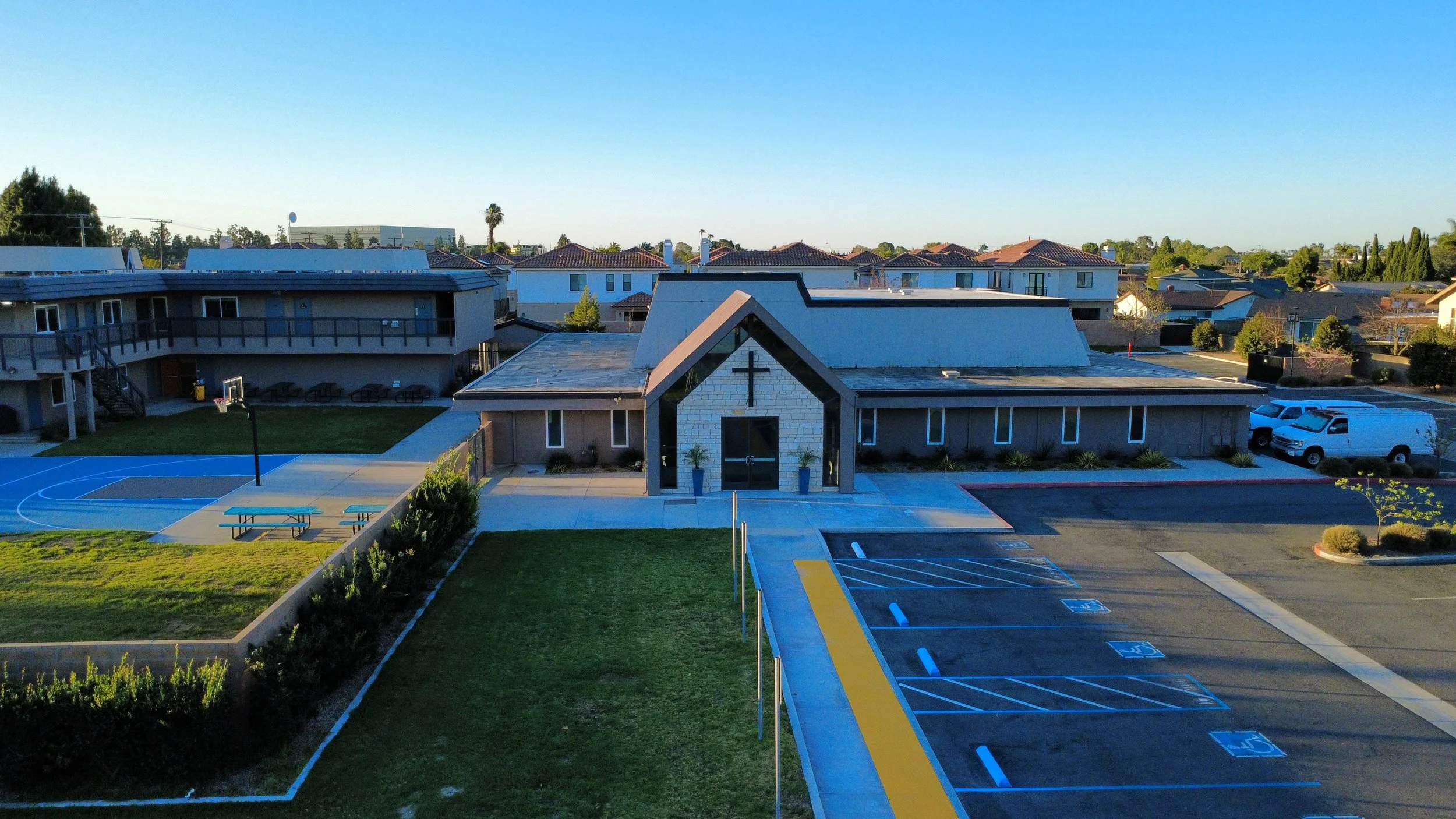 A church with a cross in front, concrete pathways, a basketball court, parking lot with designated handicapped spaces, and residential houses in the background under a clear blue sky.