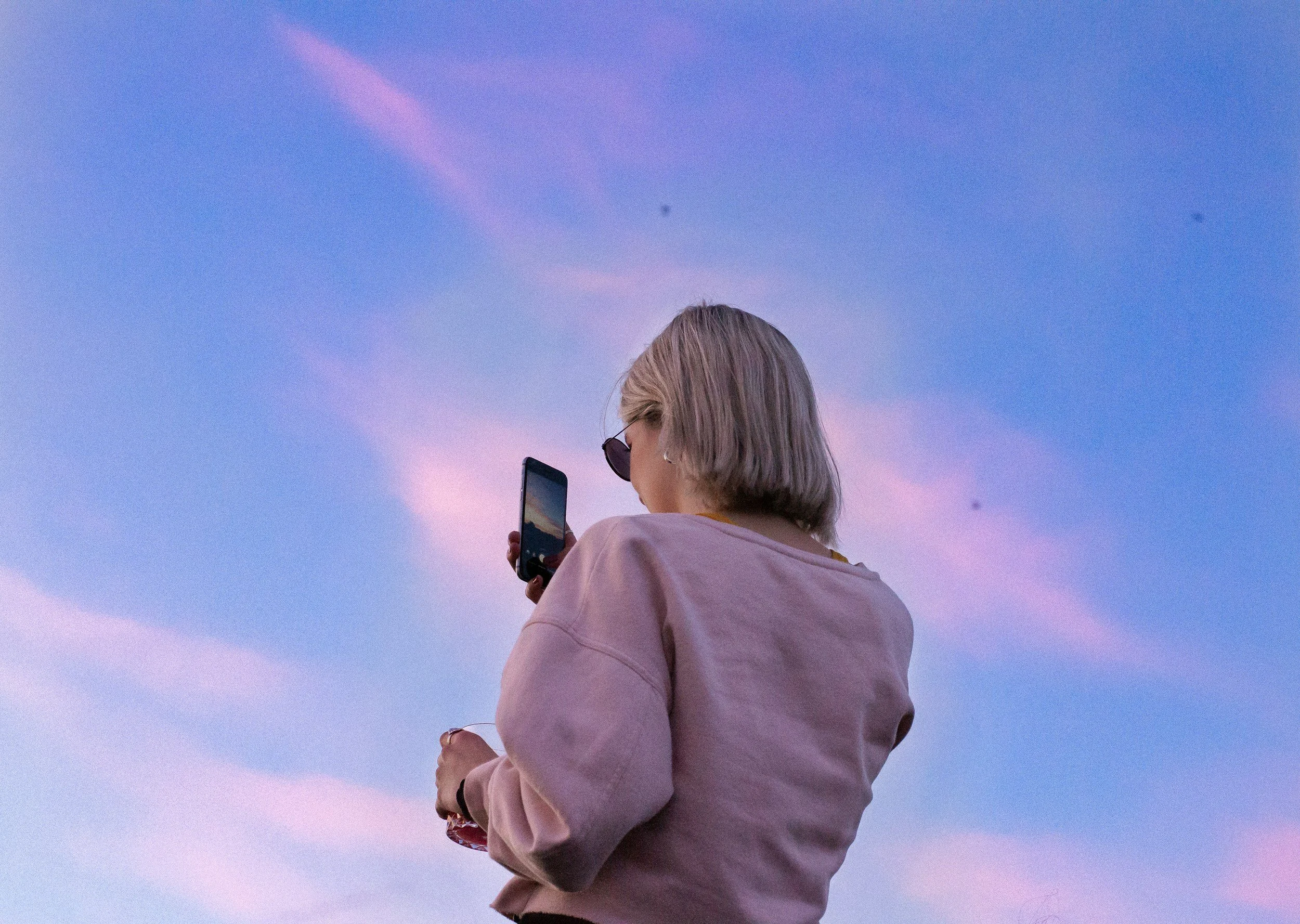 A person with blonde hair, wearing sunglasses and a light pink sweatshirt, looking at a smartphone against a sky with pink and purple hues.