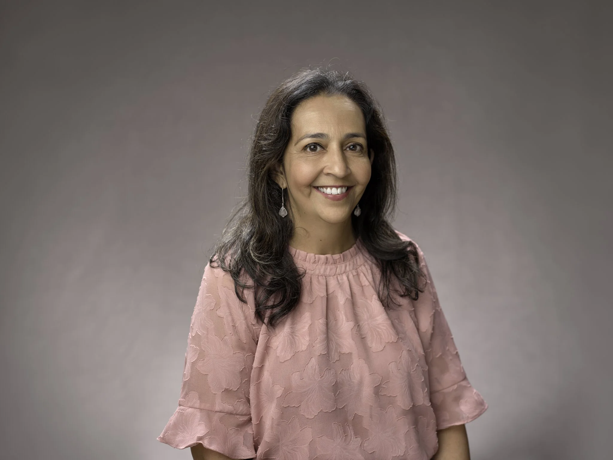 A woman with long dark hair, smiling, wearing a pink textured blouse and earrings, against a plain gray background.