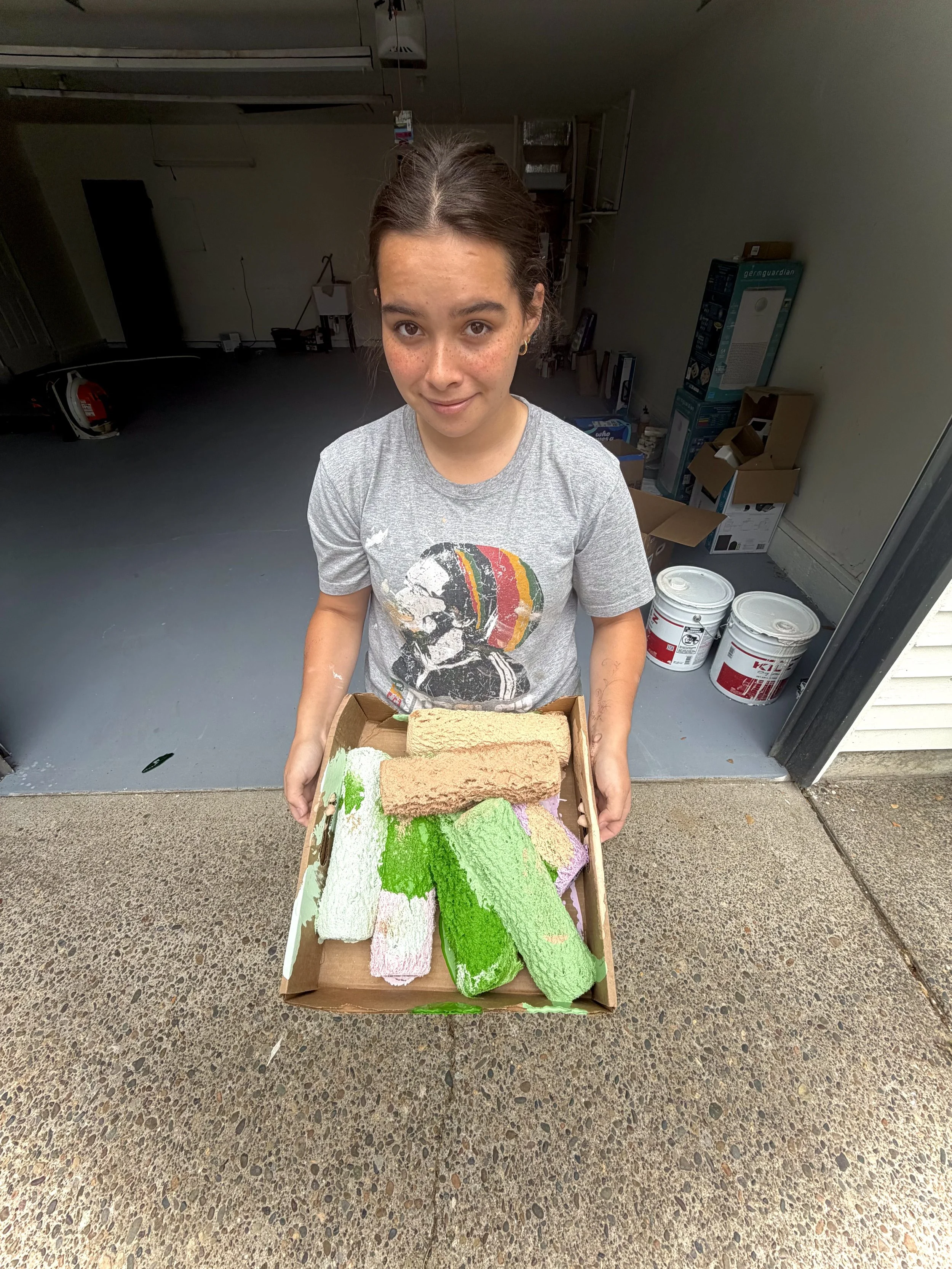 A young woman with freckles and brown hair tied back, smiling and holding a box of colorful rolled sponges in shades of green, pink, and beige. She is standing outside a garage with paint buckets and boxes visible in the background.