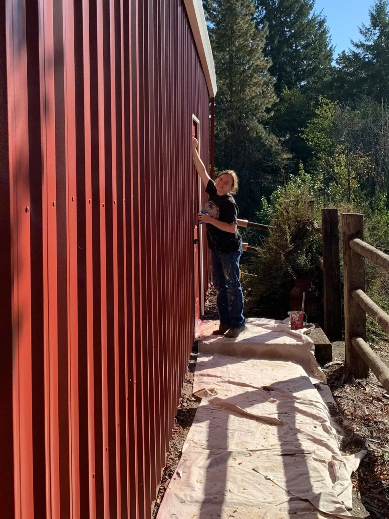 A person standing on a white tarp, painting the exterior of a red building with vertical metal siding, surrounded by trees and outdoor fencing, holding a paintbrush and a can of paint.