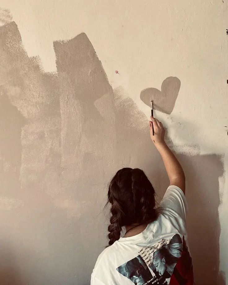 A girl with braided hair painting a gray heart on a beige wall.