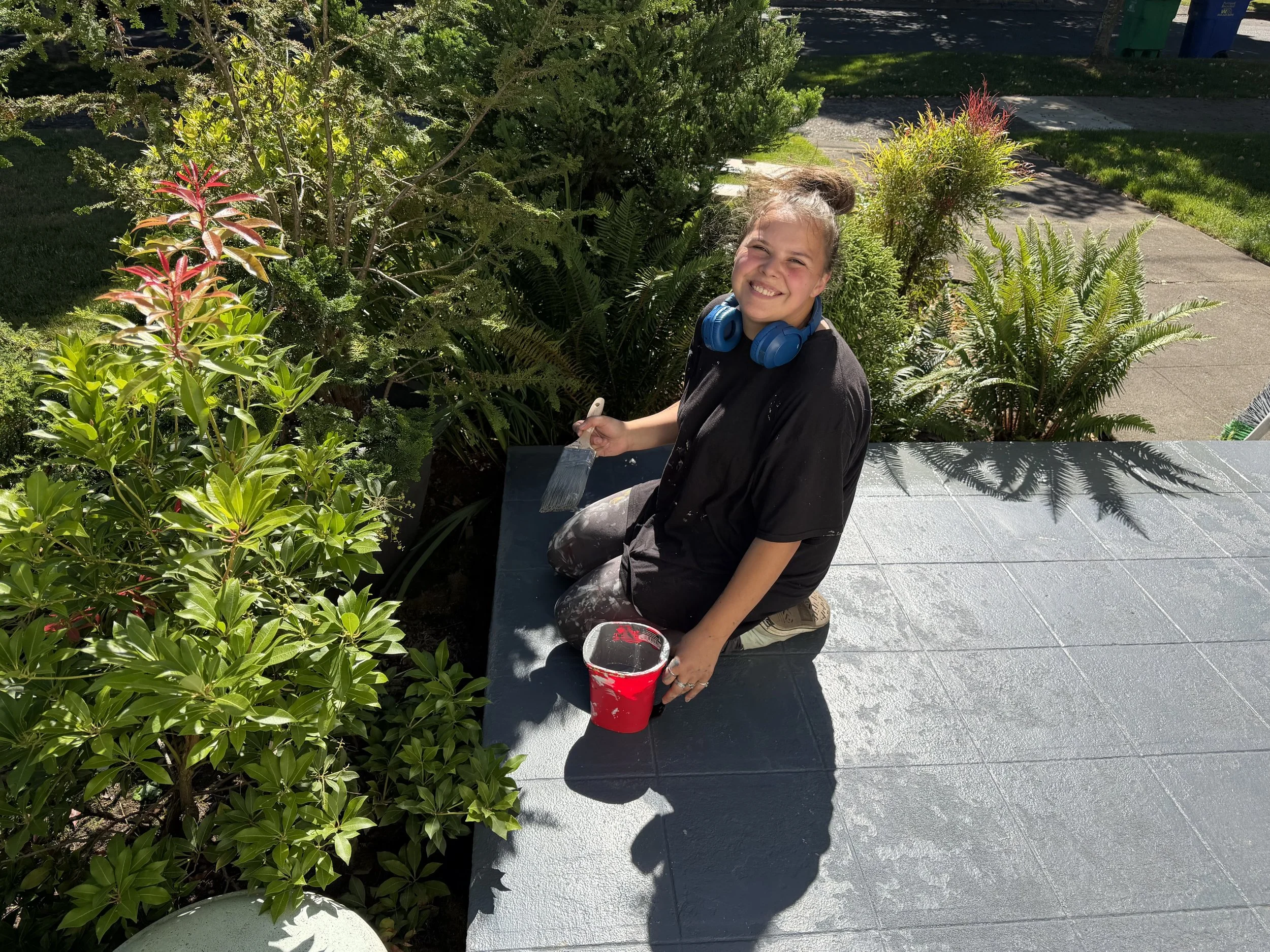 A young girl sitting on a park bench, smiling, with headphones around her neck, surrounded by lush green plants and bushes, and a sidewalk visible in the background.