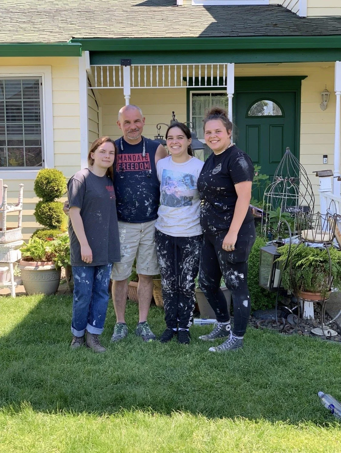 Four people standing in a front yard in front of a yellow house, with gardening plants and tools around them. They have paint splatters on their clothing and are smiling.