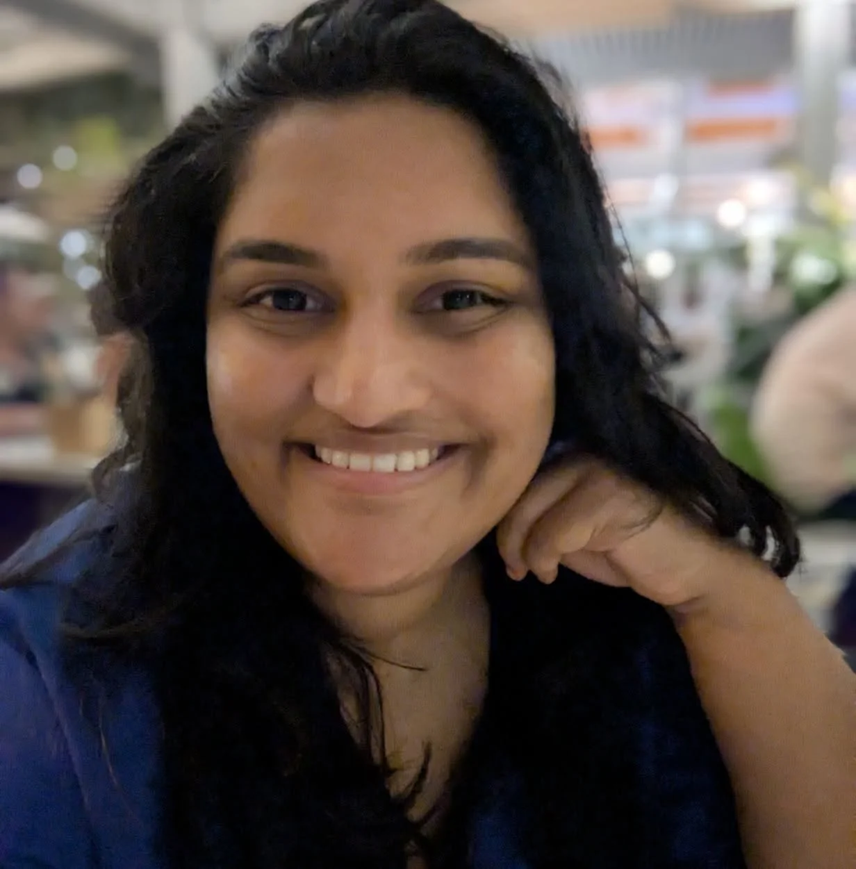 A woman smiling at the camera with her hand resting on her cheek, in a setting that appears to be a cafe or restaurant.