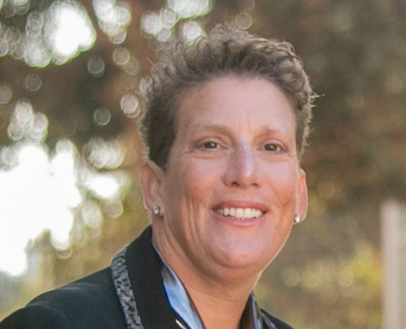 A smiling woman with short curly hair outdoors, wearing a dark blazer and pearl earrings.