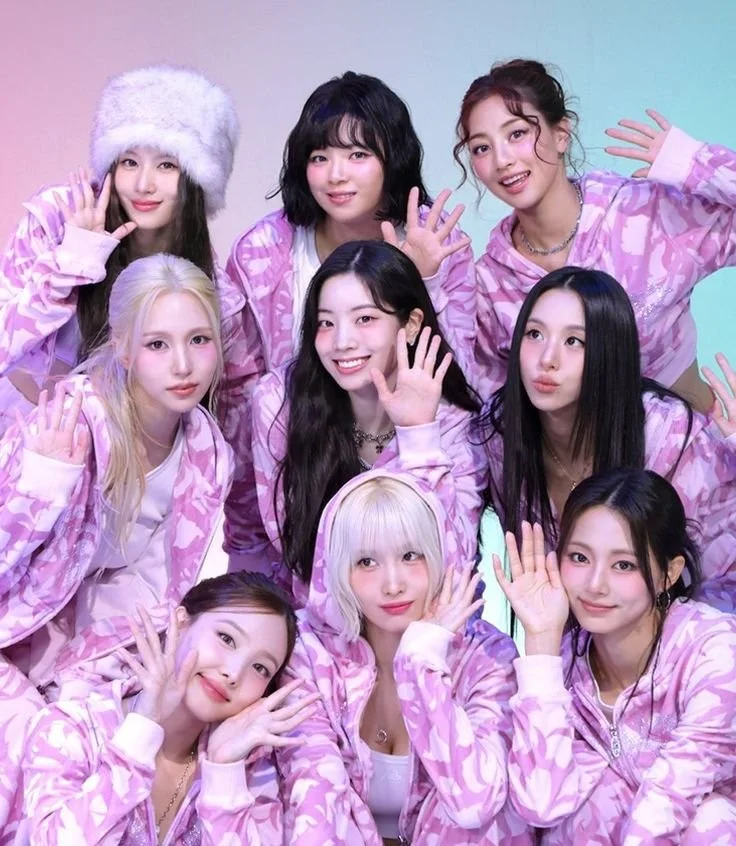 Ten young women in pink and white patterned hoodies posing together for a photo, some waving and smiling.