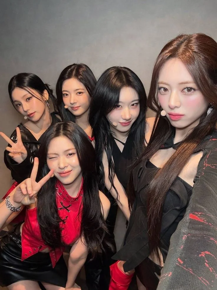 Five young women posing for a selfie, making peace signs, with a plain grey background.