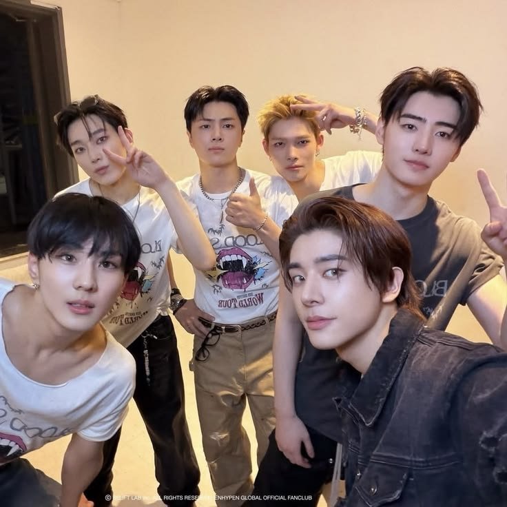 Six young men posing indoors for a group selfie, making peace signs and thumbs up gestures, with casual clothing and styled hair.