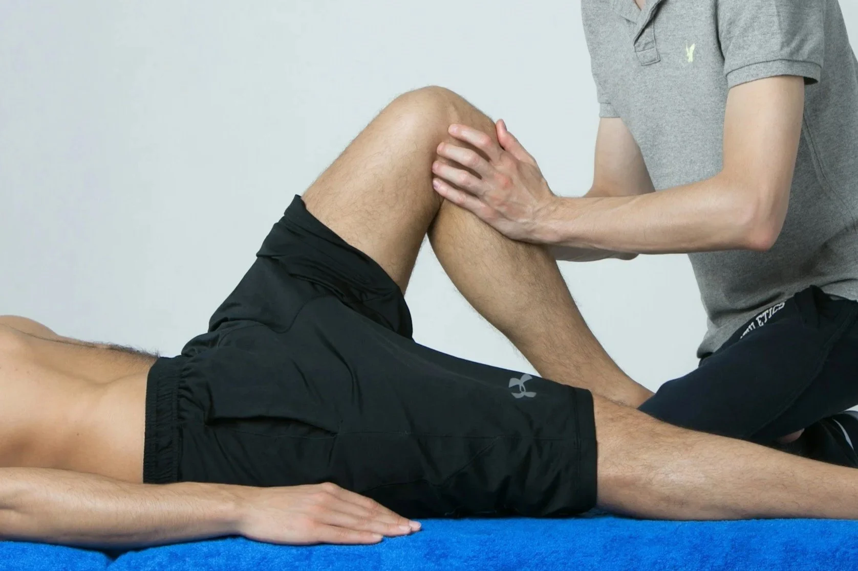 A person lying on their back on a blue mat, with another person assisting them with a knee stretch or physical therapy exercise.