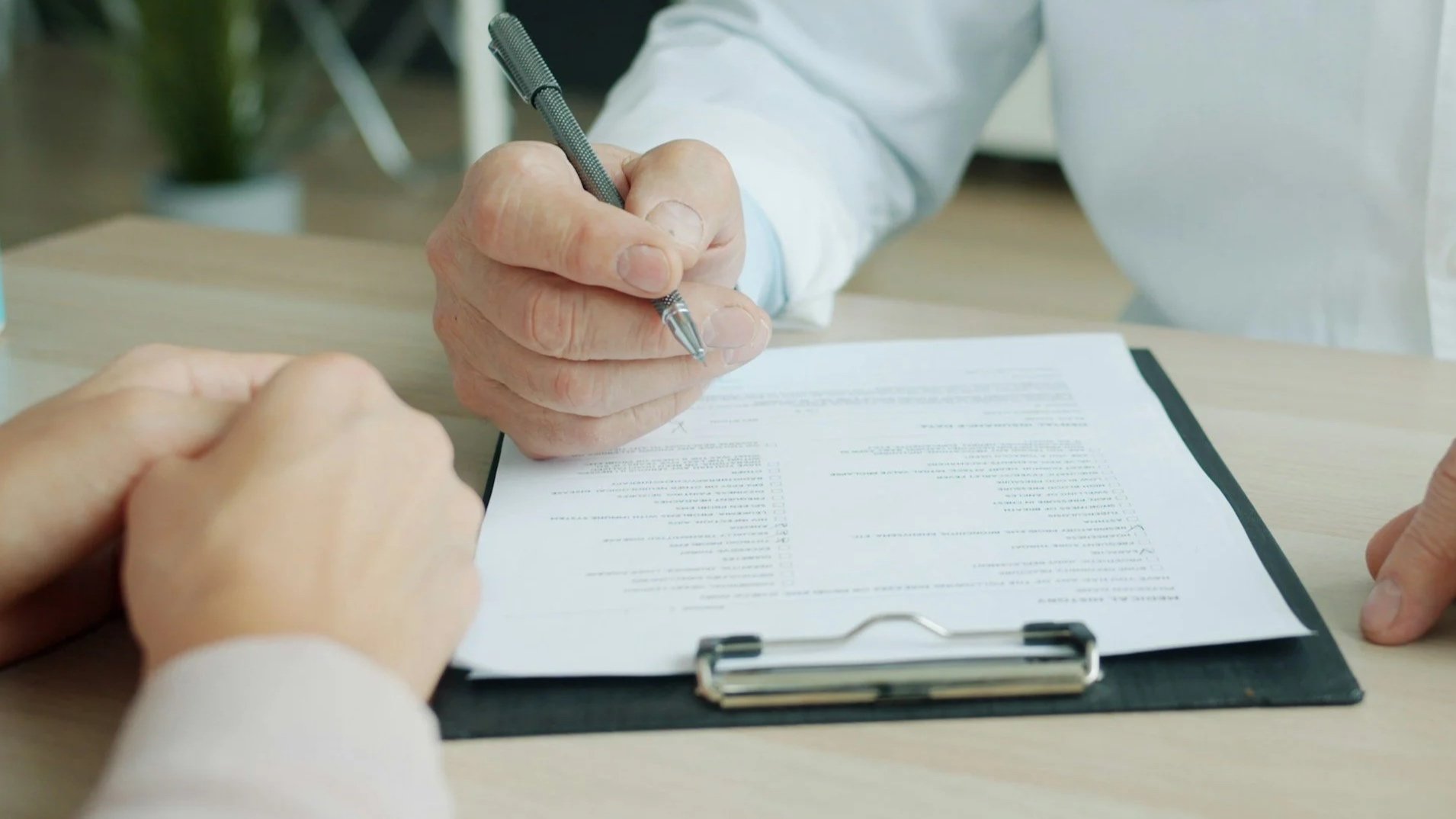 Close-up of a person's hand holding a pen over a document on a clipboard during a signing process, with another person’s hand visible.