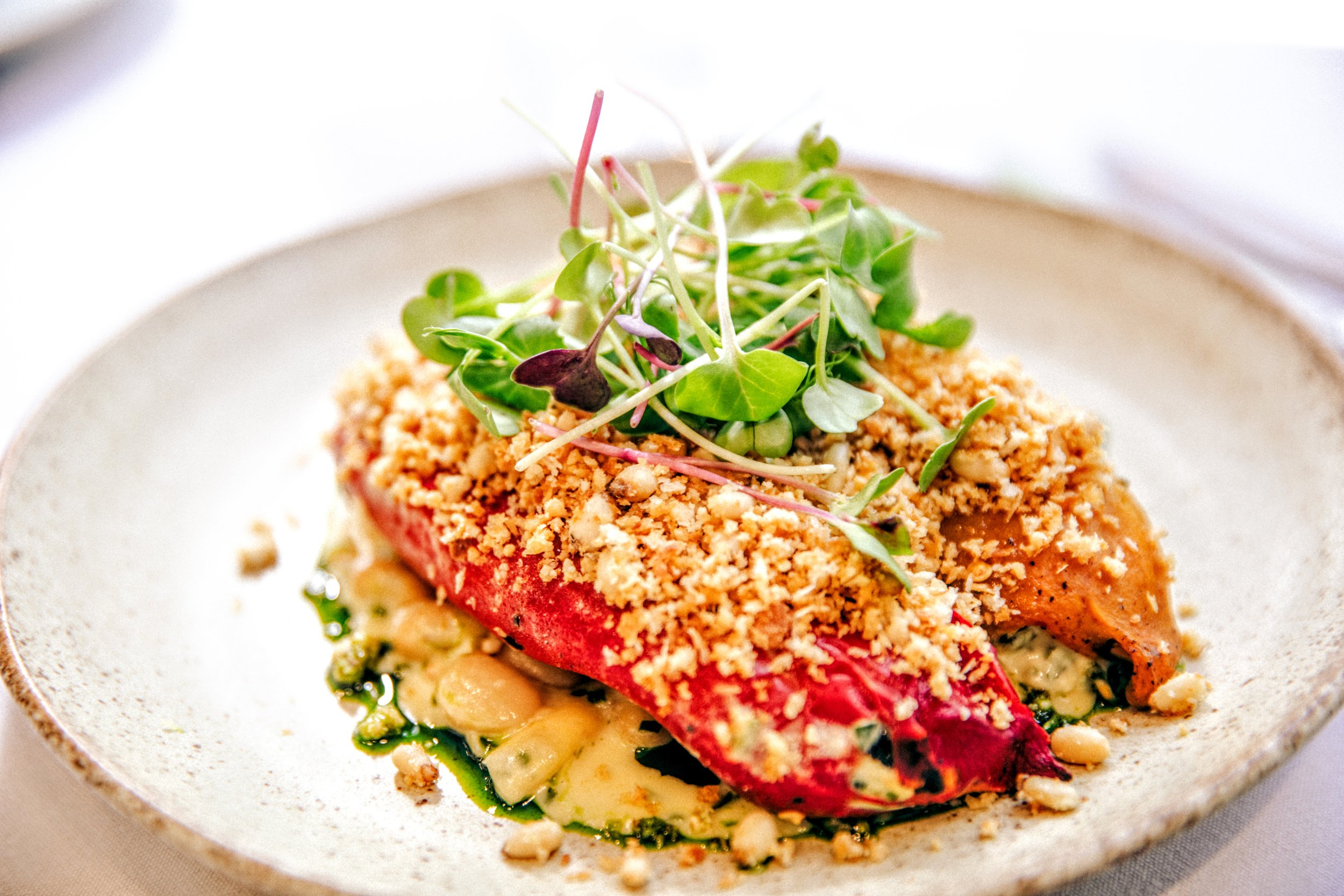 A plated dish of roasted and stuffed peppers, ricotta, lemon and spinach inside. a bed of cannellini beans with a parmesan crem sauce. pangratata and herbs on top.