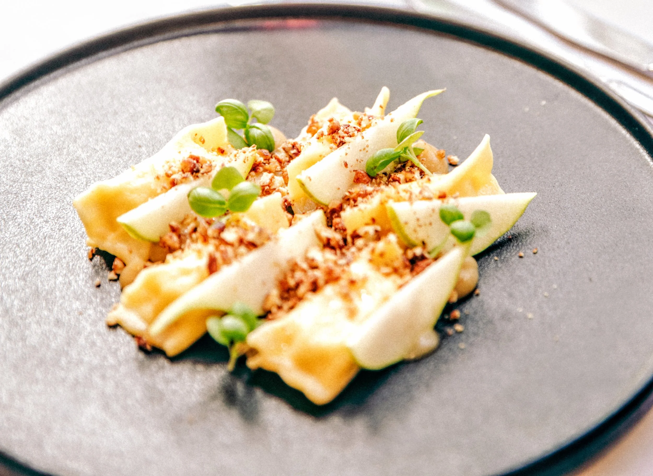 Close-up of an appetizer featuring slices of pear, blue cheese filled agnolotti, caramelized pecans, micro basil.