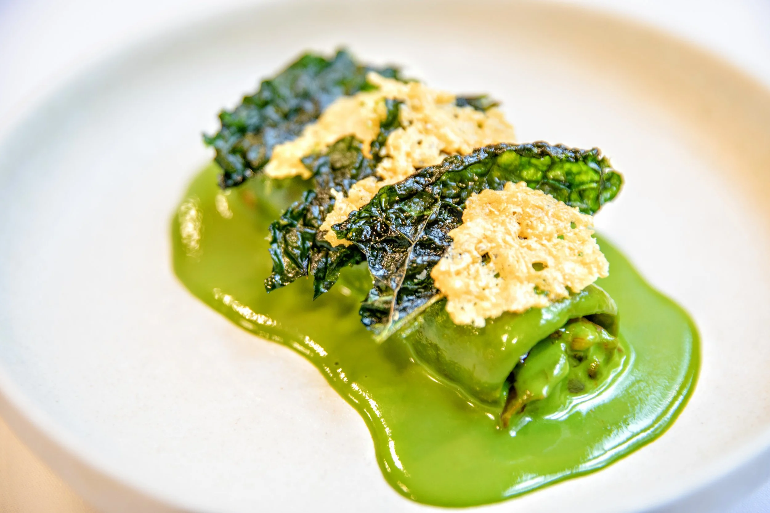 Close-up of a plated dish with gnocchi made into a sheet with peperonata stuffing and puffed pumpkon seeds, a green creamy sauce of smoked scamorza and cavolo nero, with crisp cavolo nero and parmesan crisps on top