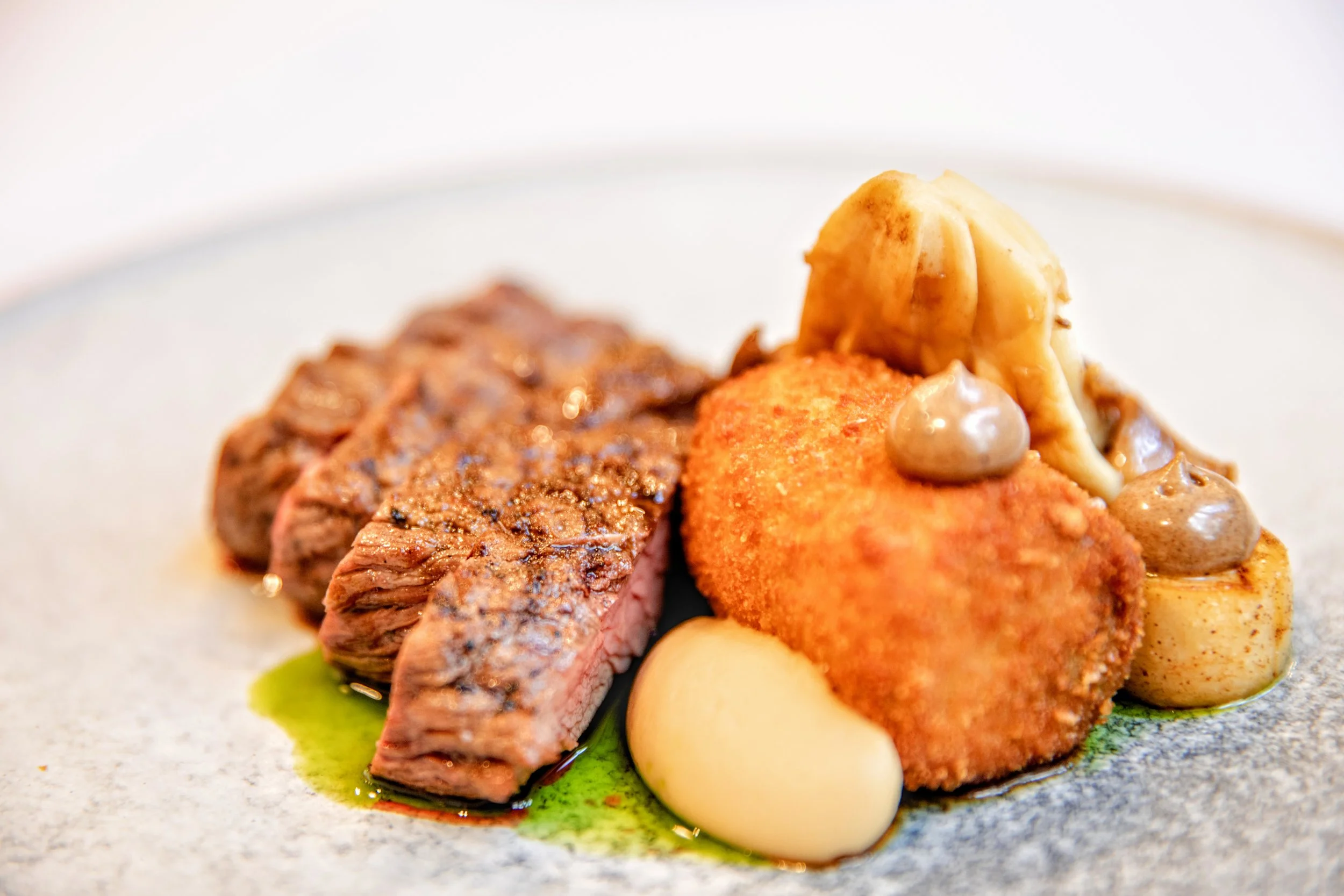 Close-up of a plated dish featuring sliced barbequed beef bavette steak, a croquette of beef chuck, ewes cheese ad mushrooms. oyster mushrooms, a black garlic mayonnaise, celeriac puree, celeriac fondants and a red wine jus.
