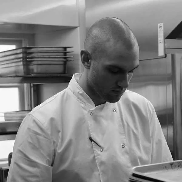 Chef James Welch in chef whites, focused, in a professional kitchen.
