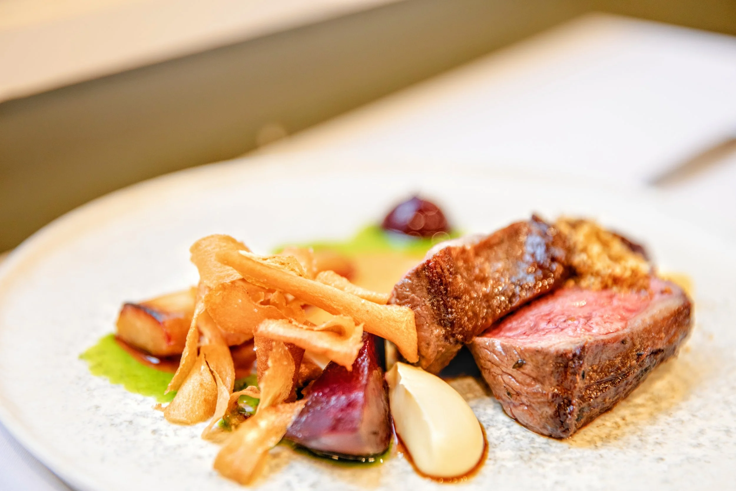 Close-up of a plated gourmet meal featuring rare venison picanha, fermented blueberry 'ketchup' parsnips 3 ways, dukkha, smoked beetroot and a red wine sauce.