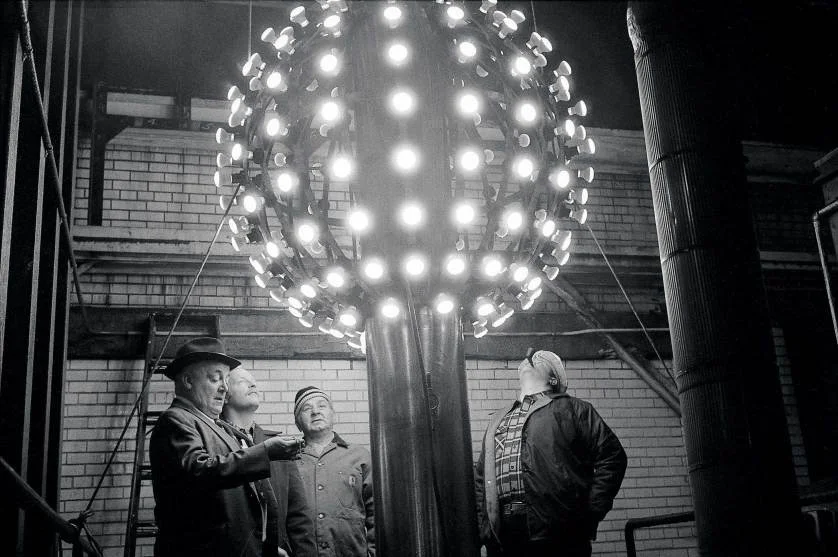 Four men standing around a large illuminated the New Years Eve times square mast in New York City  in an industrial setting with brick walls and pipes.
