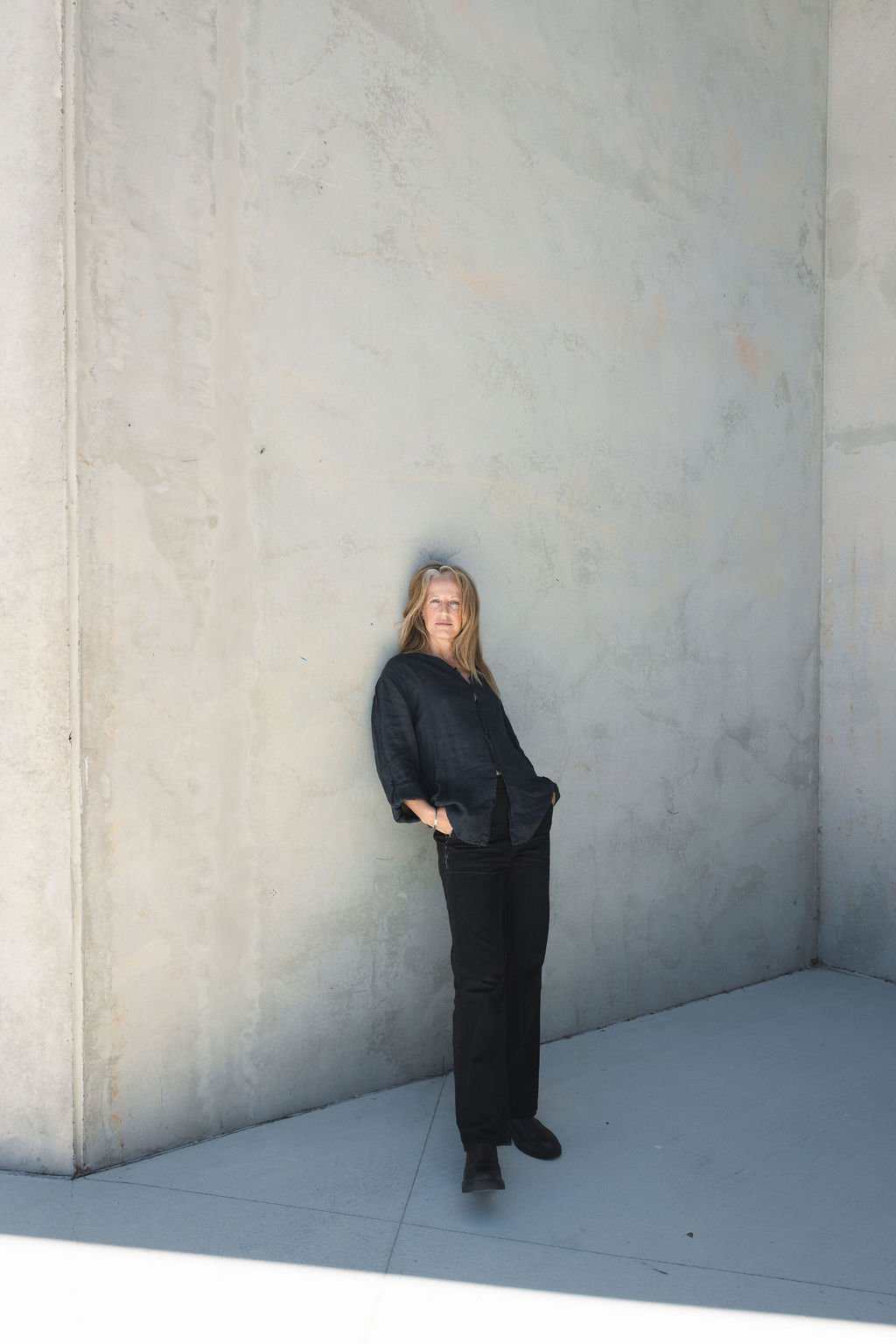 Woman in black jacket and pants standing against a concrete wall with hands in pockets.