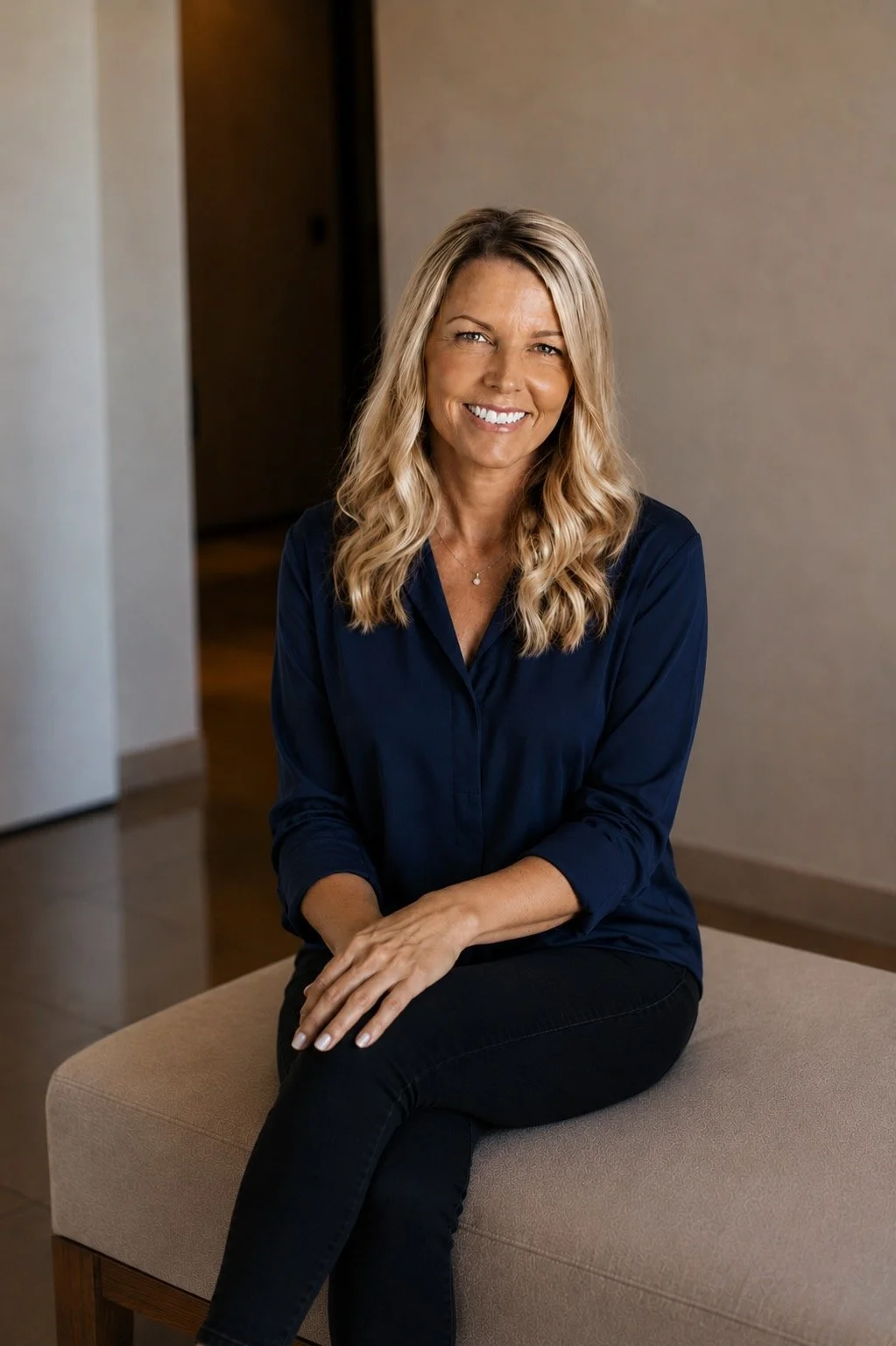 A woman with blonde wavy hair, smiling, sitting on a beige upholstered bench in a softly lit indoor setting, wearing a navy blue long-sleeve blouse and black pants.