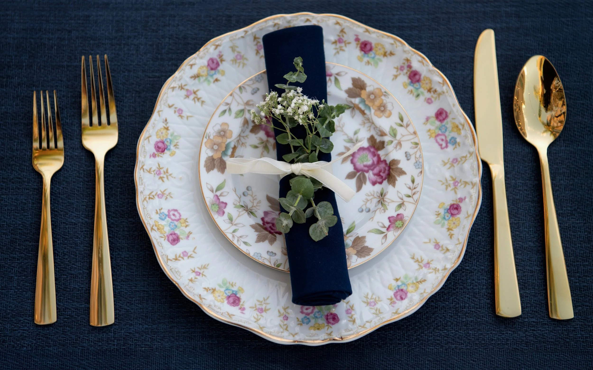 Elegant place setting with floral patterned dinner plate, dark blue napkin, small bouquet of white flowers and greenery, gold utensils, on dark blue tablecloth.