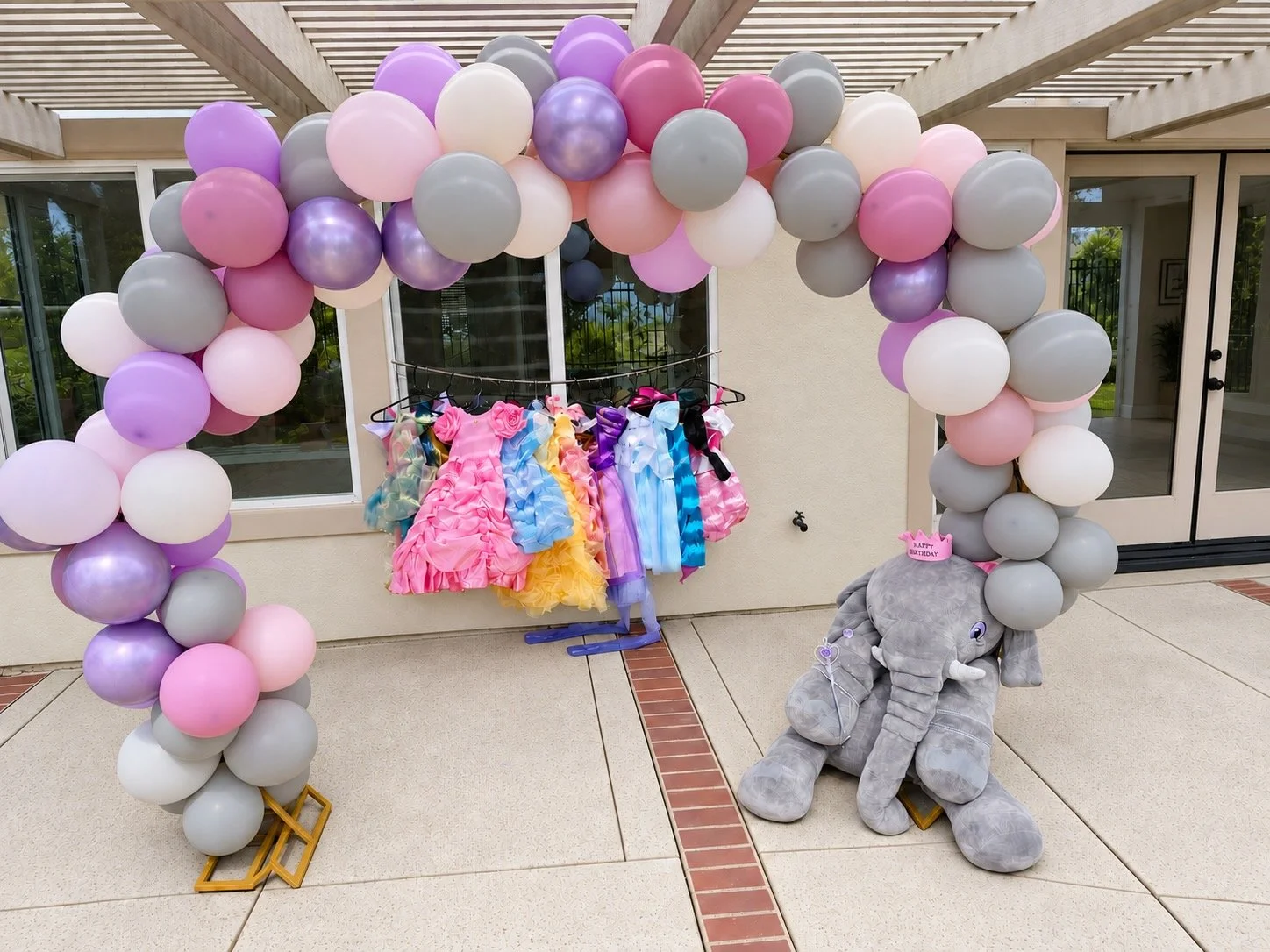 Decorative setup with a pink, purple, white, and gray balloon arch, a rack of colorful dresses, and a stuffed gray elephant toy with a pink birthday hat marked 'Happy Birthday'.
