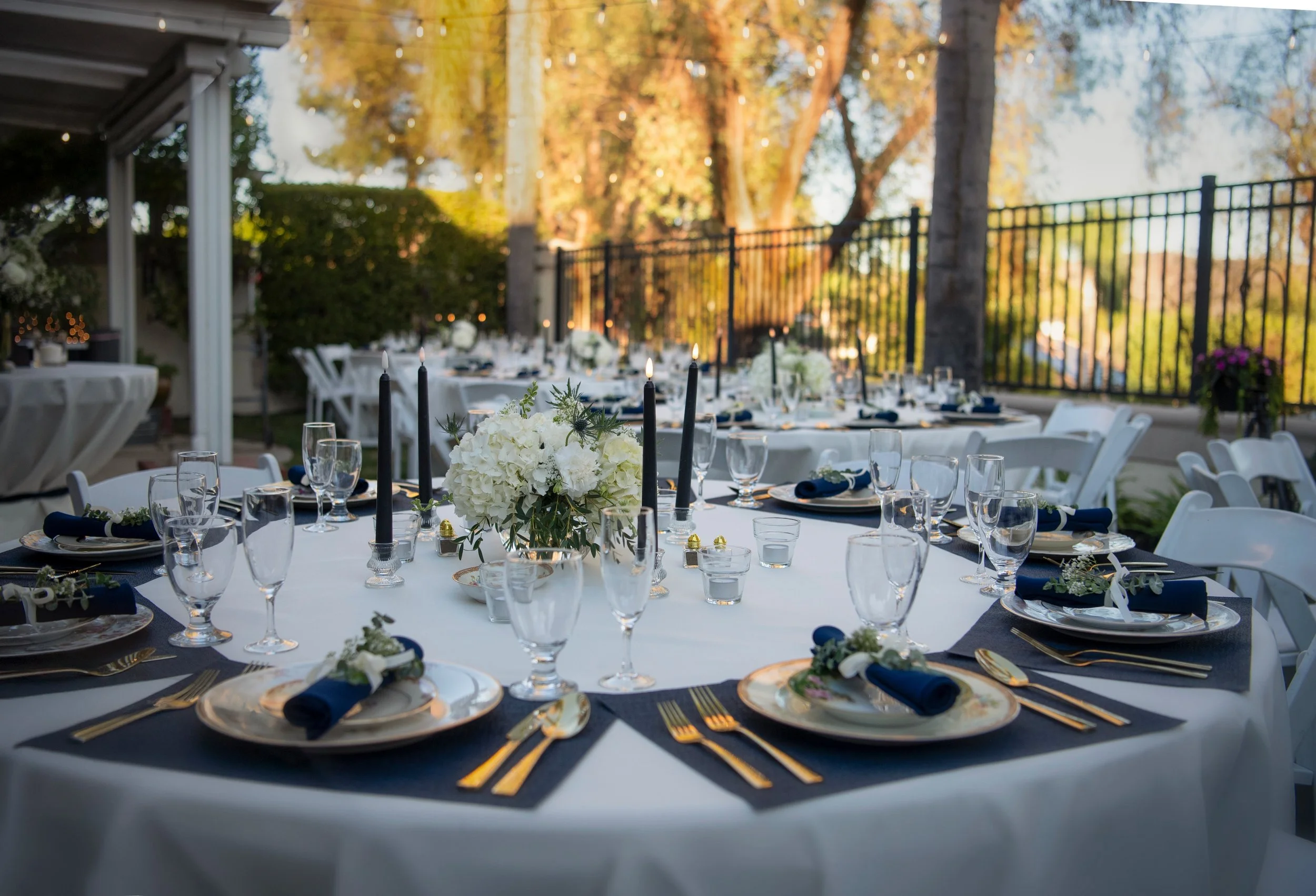 Elegant outdoor banquet table set for a formal event with white tablecloths, floral centerpieces, blue napkins, gold cutlery, and black candles, with a backdrop of trees and string lights in a garden setting during golden hour.