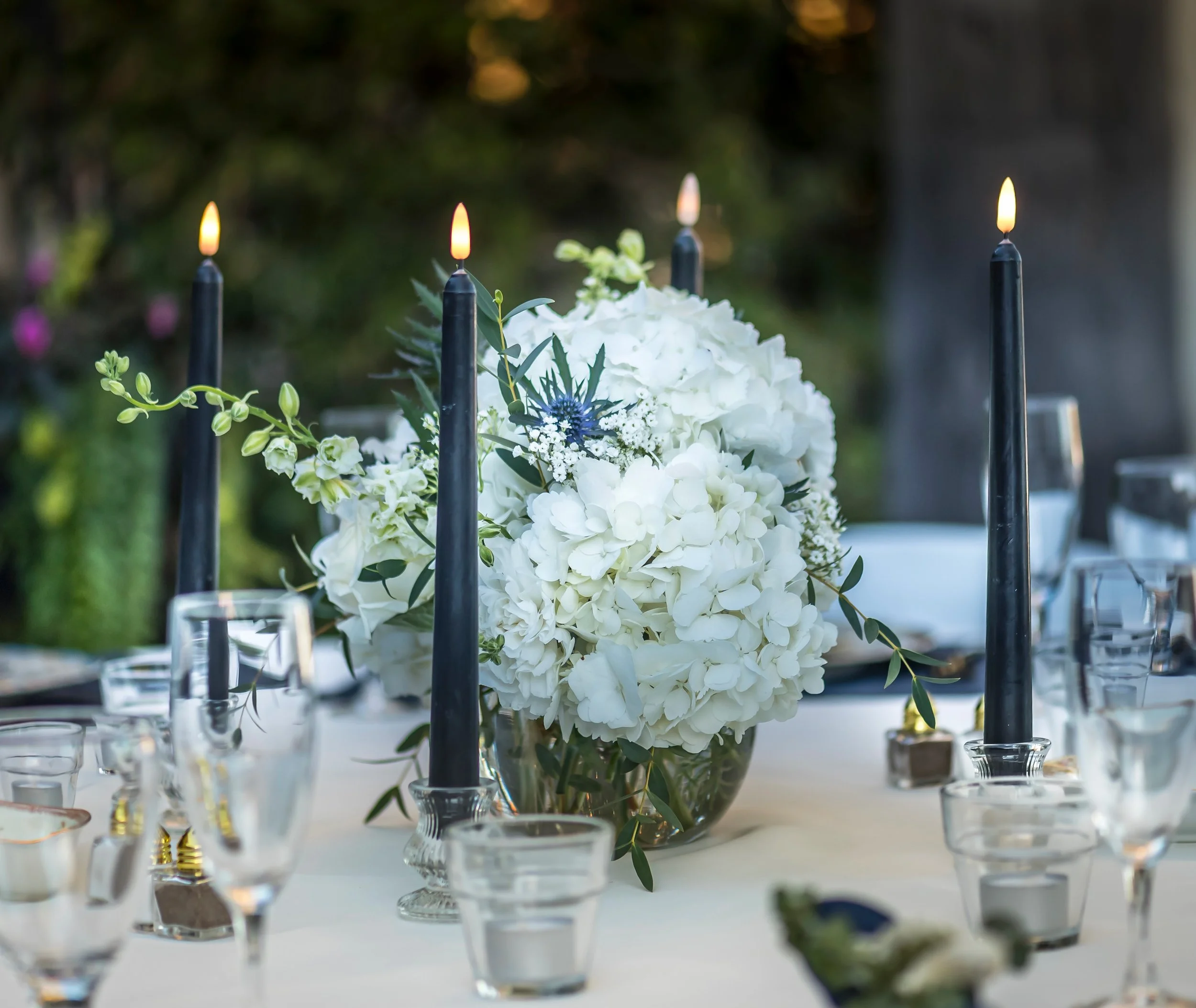 A floral centerpiece with white hydrangeas and greenery, surrounded by lit black taper candles, arranged on a white table with glassware.
