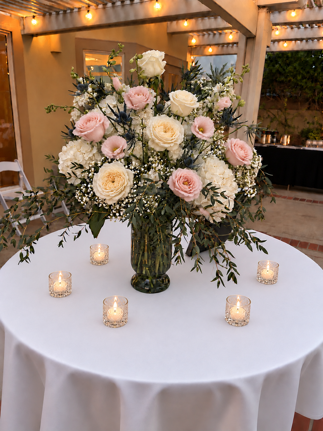 A large floral centerpiece on a round table with a white tablecloth, surrounded by five lit candles in glass holders. The arrangement includes white and pale pink roses, white hydrangeas, and greenery. The setting appears to be an outdoor or semi-outdoor event space with warm lighting and wooden beams overhead.