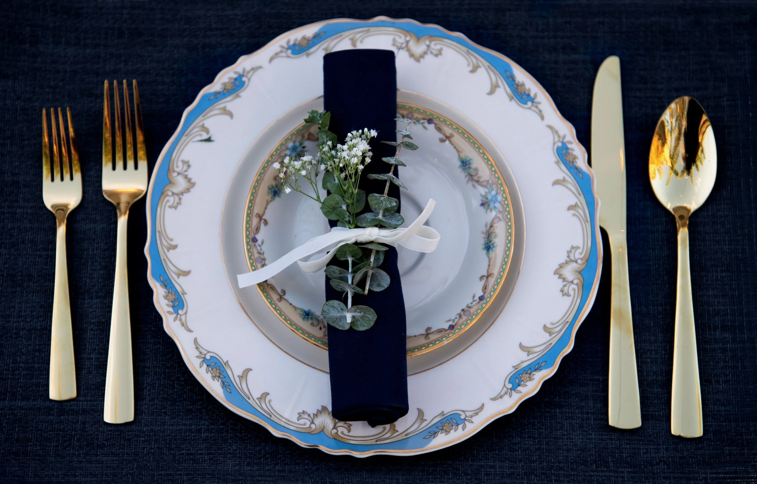 Elegant table setting with a decorative blue and gold trimmed plate, gold-colored cutlery, a rolled black napkin tied with a cream ribbon, and a small floral arrangement with greenery on a dark tablecloth.