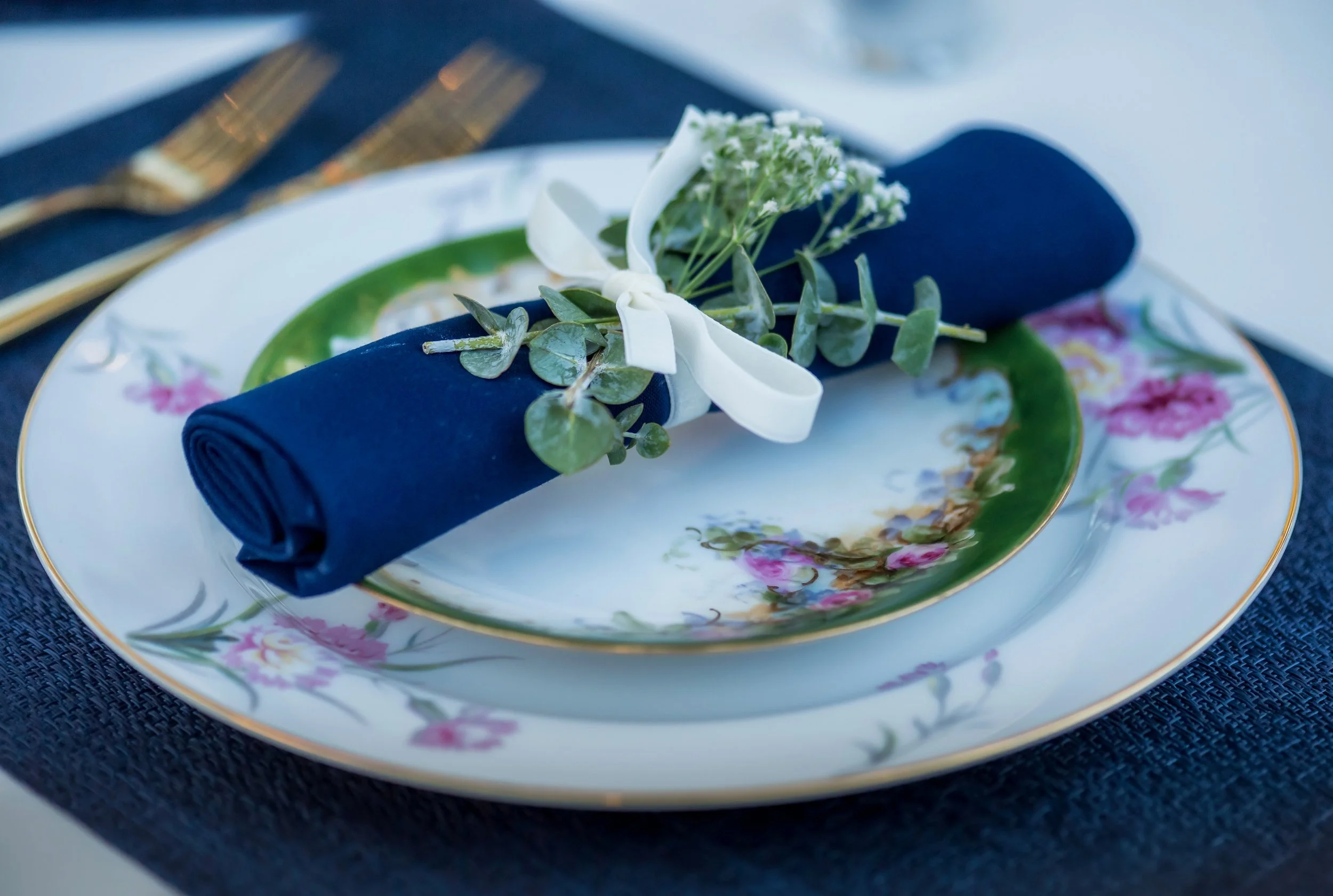 Set table with floral-patterned china plate, navy blue napkin wrapped with a small bouquet of white flowers and greenery, gold-toned cutlery, on a dark textured placemat.