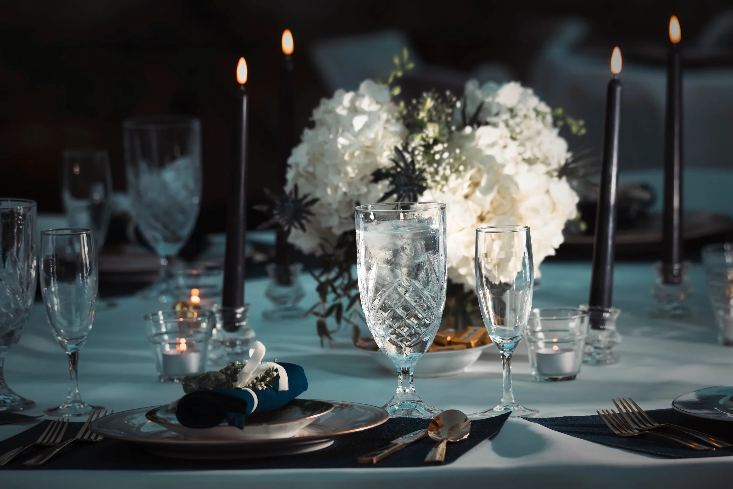 Elegant dinner table setting with glassware, floral centerpiece, candles, and plated utensils in a dimly lit environment.