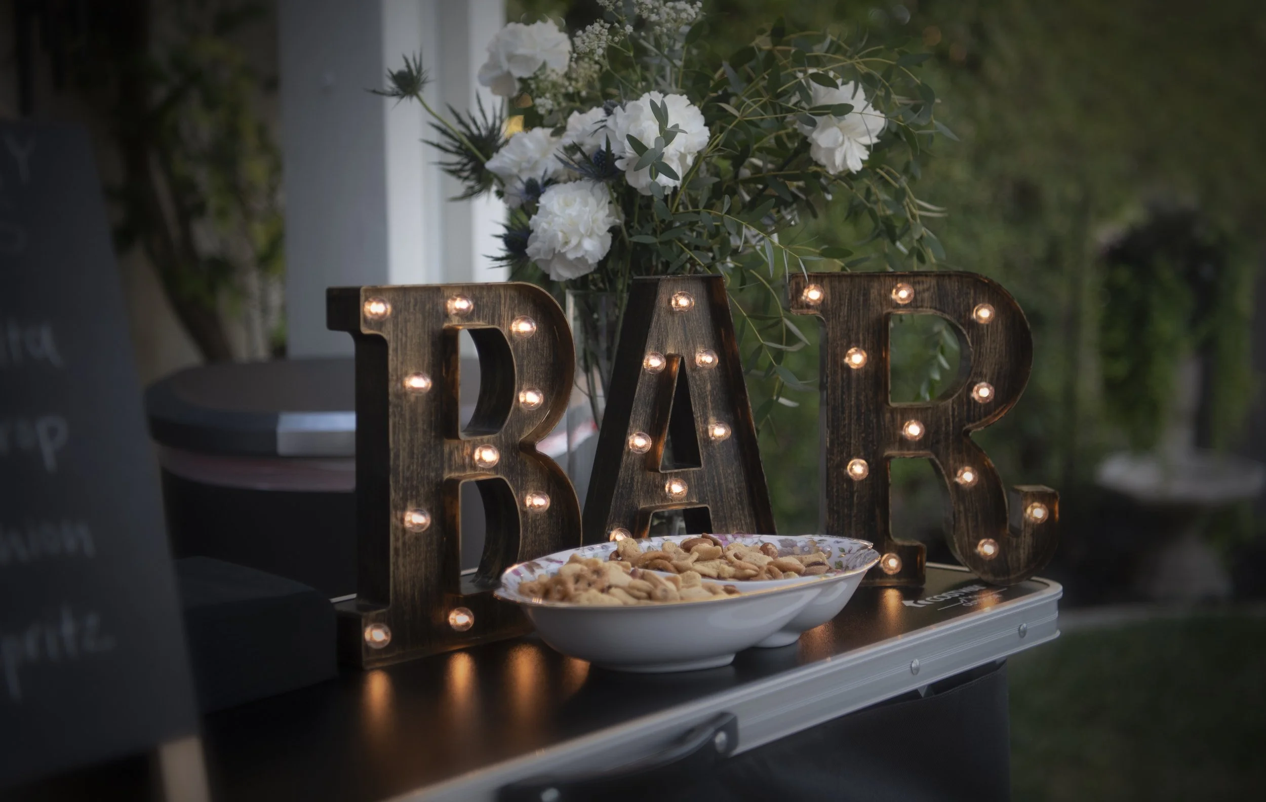 Decorative lit sign spelling 'BAR' surrounded by a bouquet of white flowers and greenery, with a dish of snacks in the foreground, set outdoors during evening.