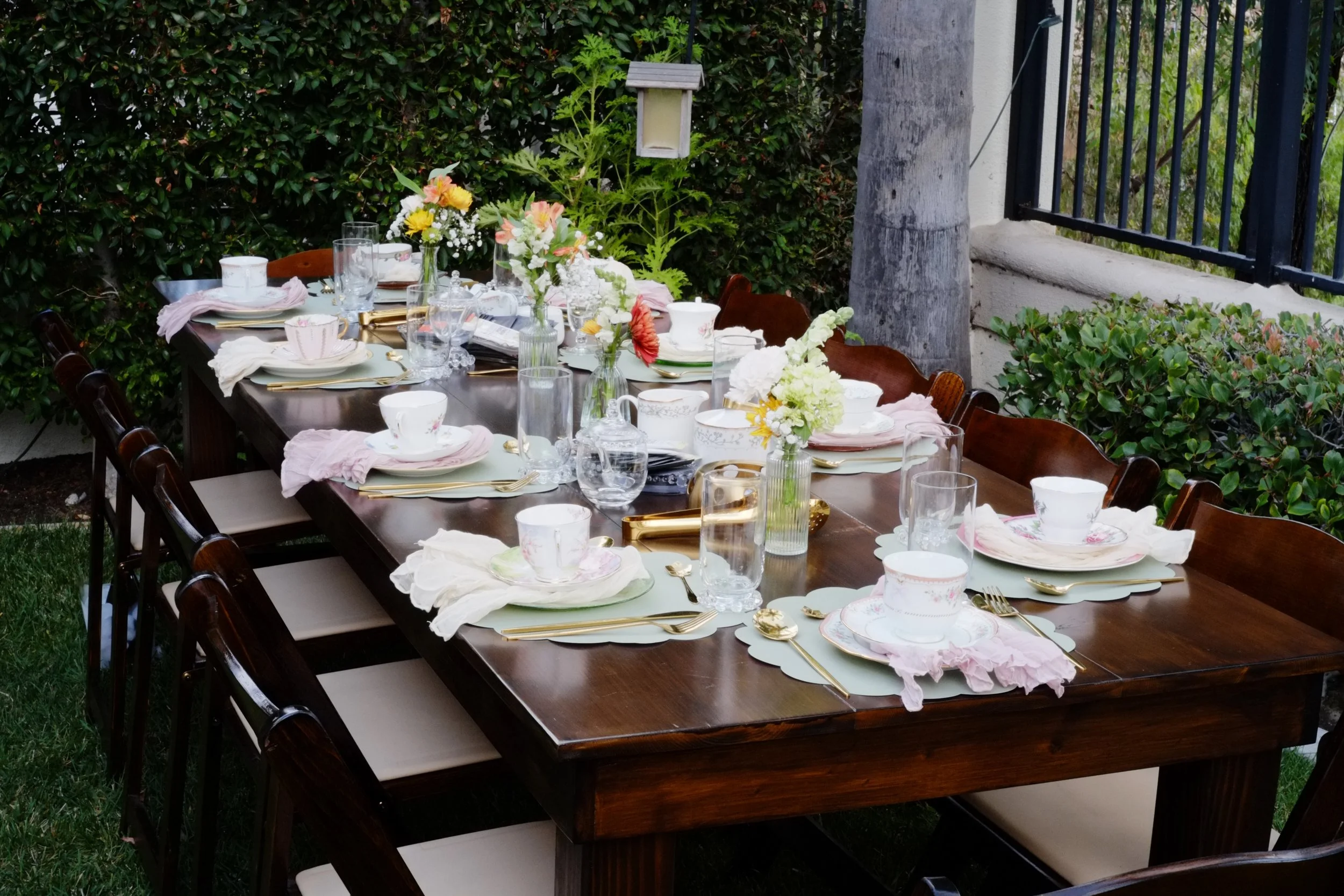 A long wooden outdoor dining table set for a meal with floral centerpieces, arranged with plates, cups, glasses, and gold utensils, surrounded by chairs, with lush greenery and a black iron fence in the background.