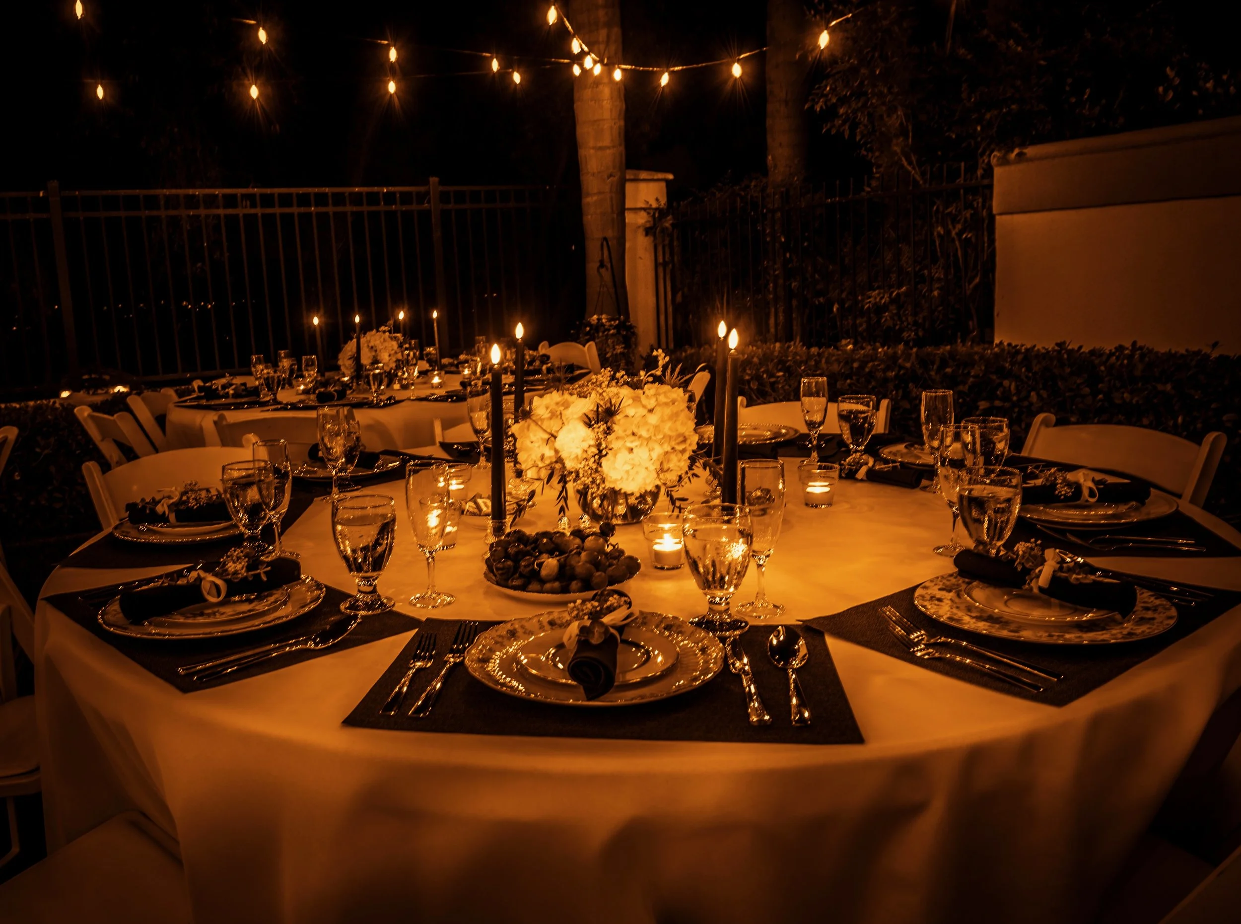 Nighttime outdoor dinner setup with a round table, decorated with flowers, candles, and glassware, illuminated by string lights and surrounded by a metal fence, trees, and plants.