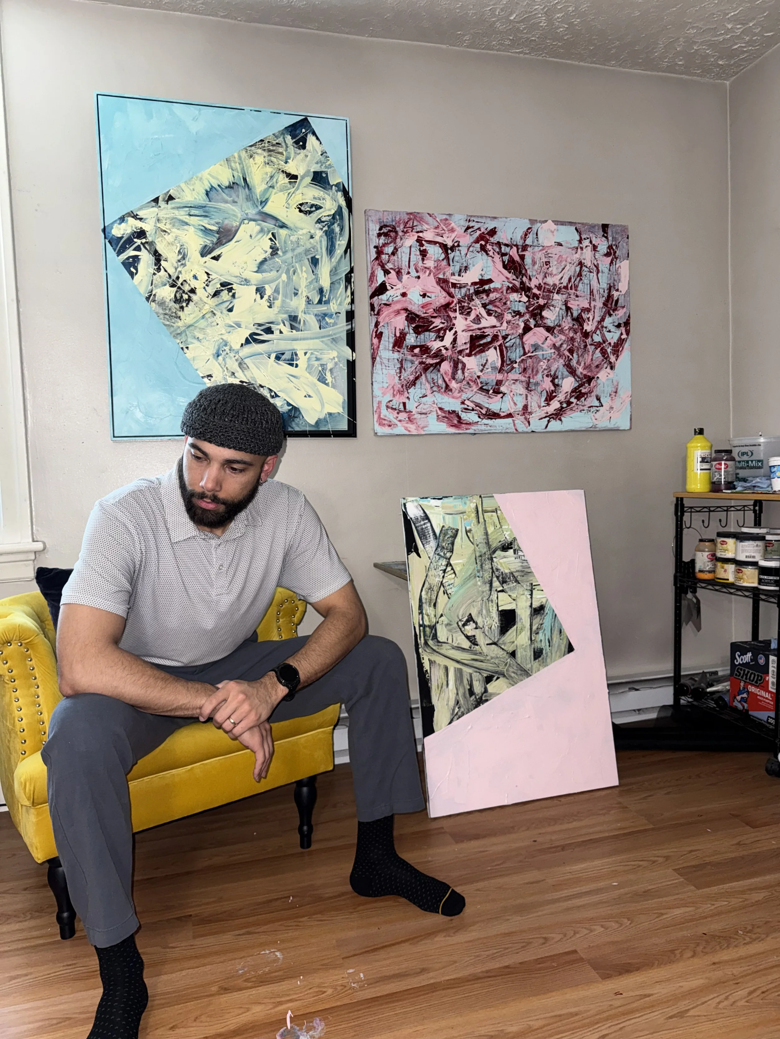 A man wearing a gray shirt, gray pants, black socks, and a black knit cap is sitting on a yellow velvet armchair with black wooden legs in an art studio. Behind him, there are several abstract paintings on the wall and leaning against the wall. To the right, a black metal shelf holds art supplies, including yellow and white containers.
