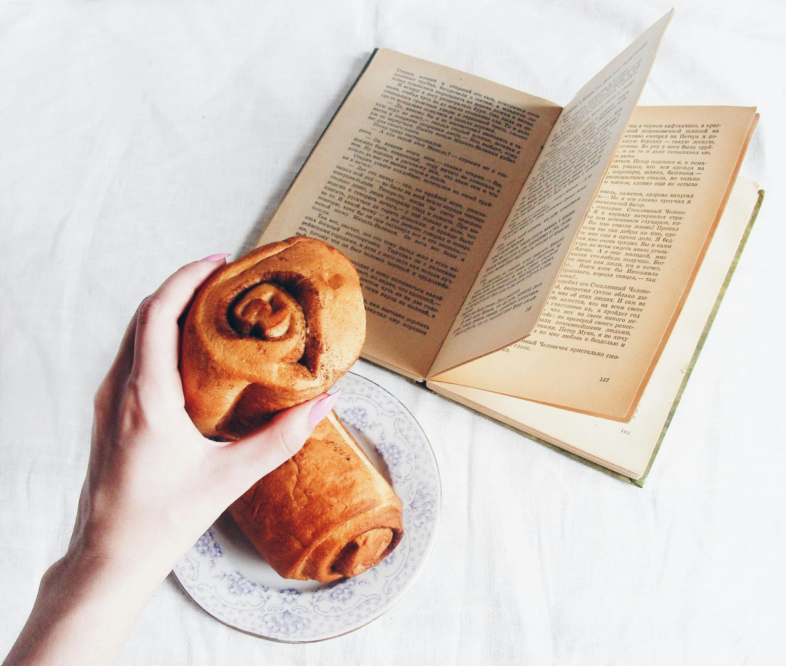 A hand holding a cinnamon roll on a white patterned plate, with an open book in the background on a white surface.