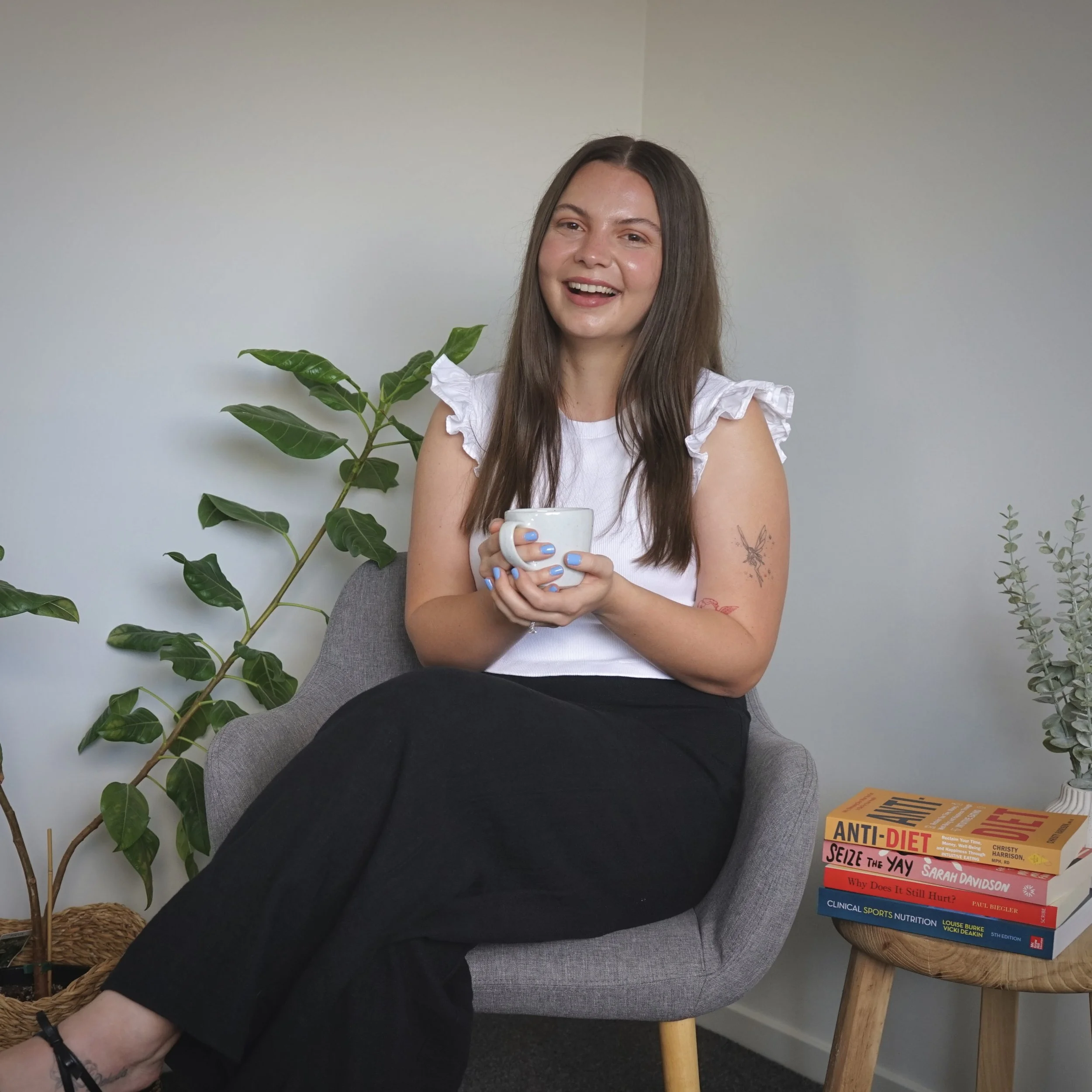 A woman with long brown hair smiling and holding a white mug, seated on a gray chair in front of a plain wall, with a large green leafy plant to her left and a stack of health and diet books on a wooden side table to her right.