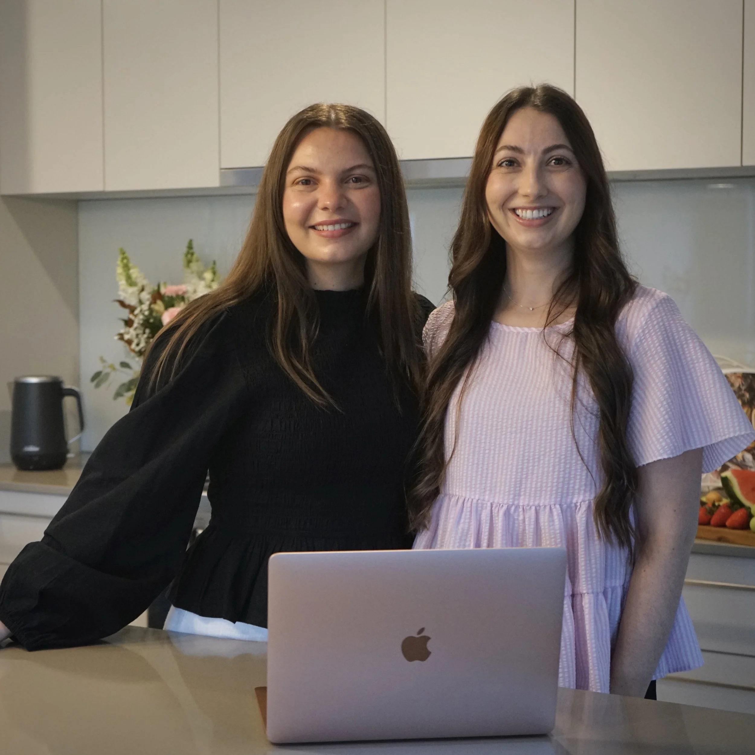 Two women stand behind a desk with a laptop, smiling at the camera in a kitchen or dining area with a vase of flowers in the background.