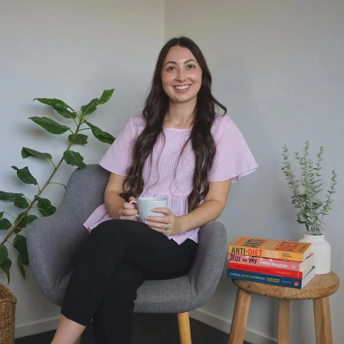 A woman with long wavy brown hair smiling and holding a white mug, sitting in a gray armchair next to a small wooden side table with books and a plant, in a cozy room.