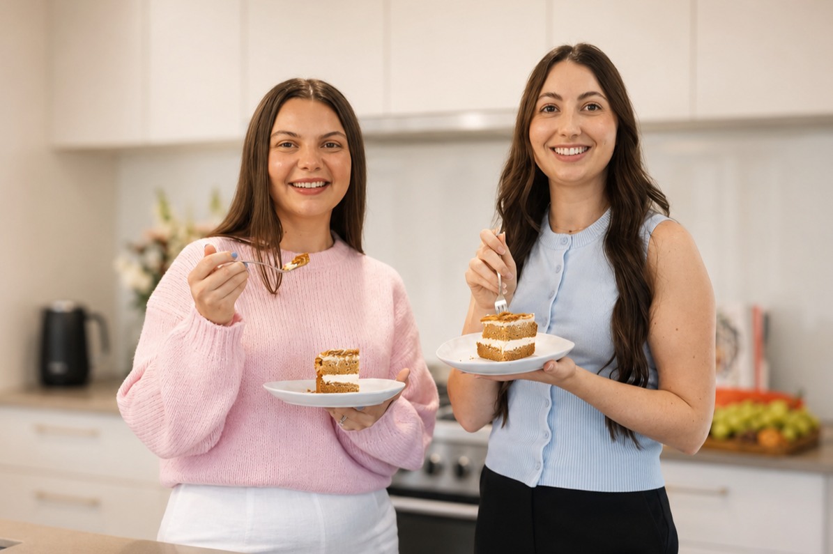 Two women smiling in a kitchen, each holding a plate with a slice of layered cake and a fork.