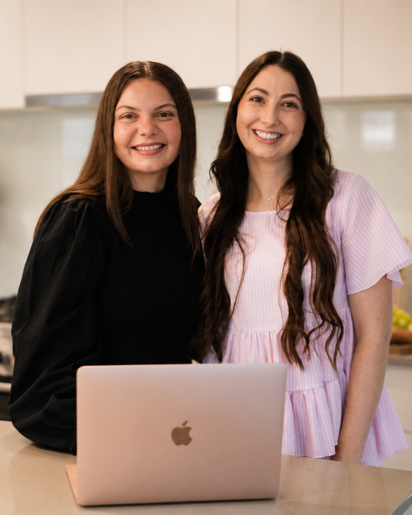 Two women standing in a kitchen behind a pink MacBook, smiling at the camera.