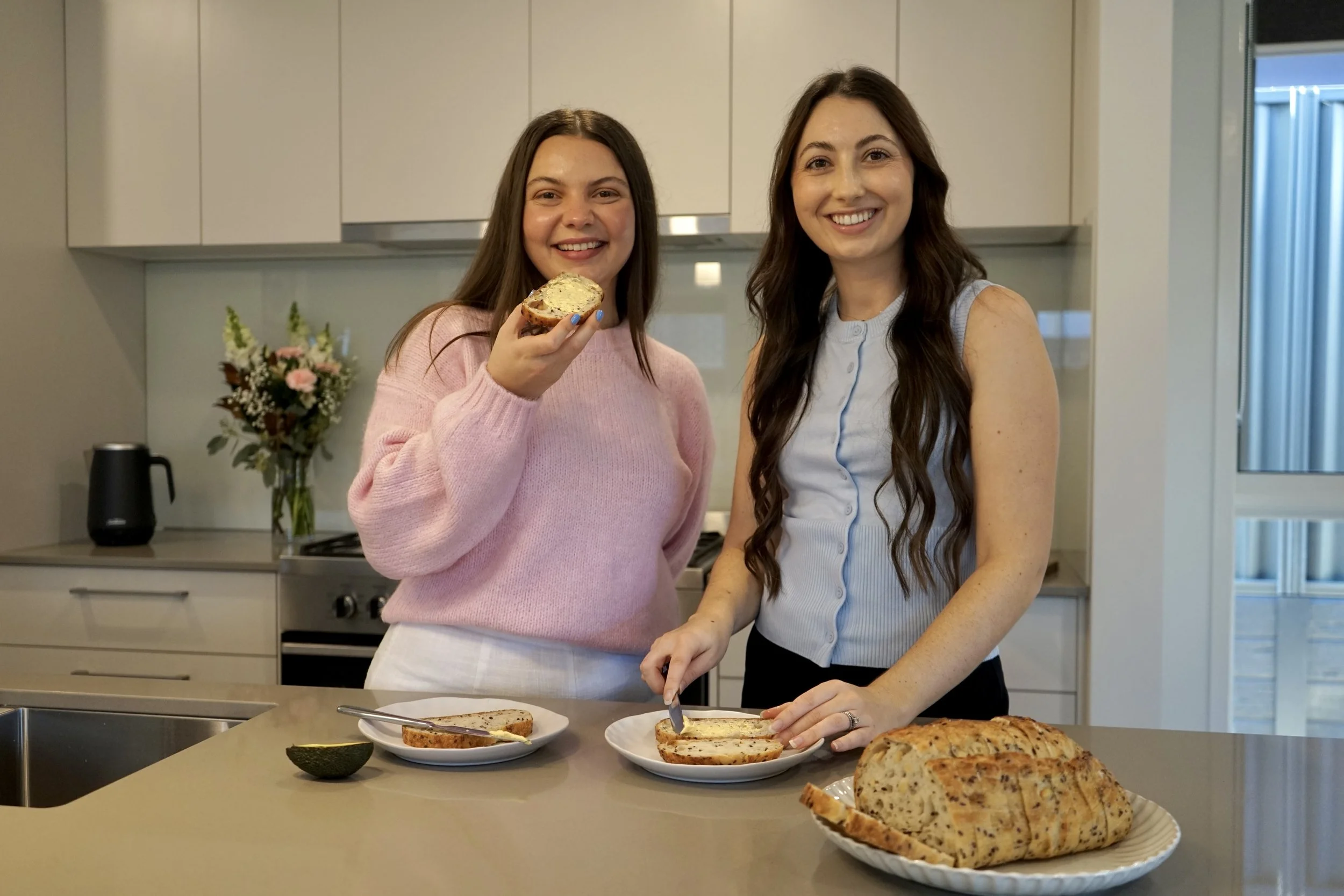 Two women smiling in a kitchen, preparing bread and spread on slices, with a loaf of bread on a plate in the foreground and plates of bread slices on the counter.