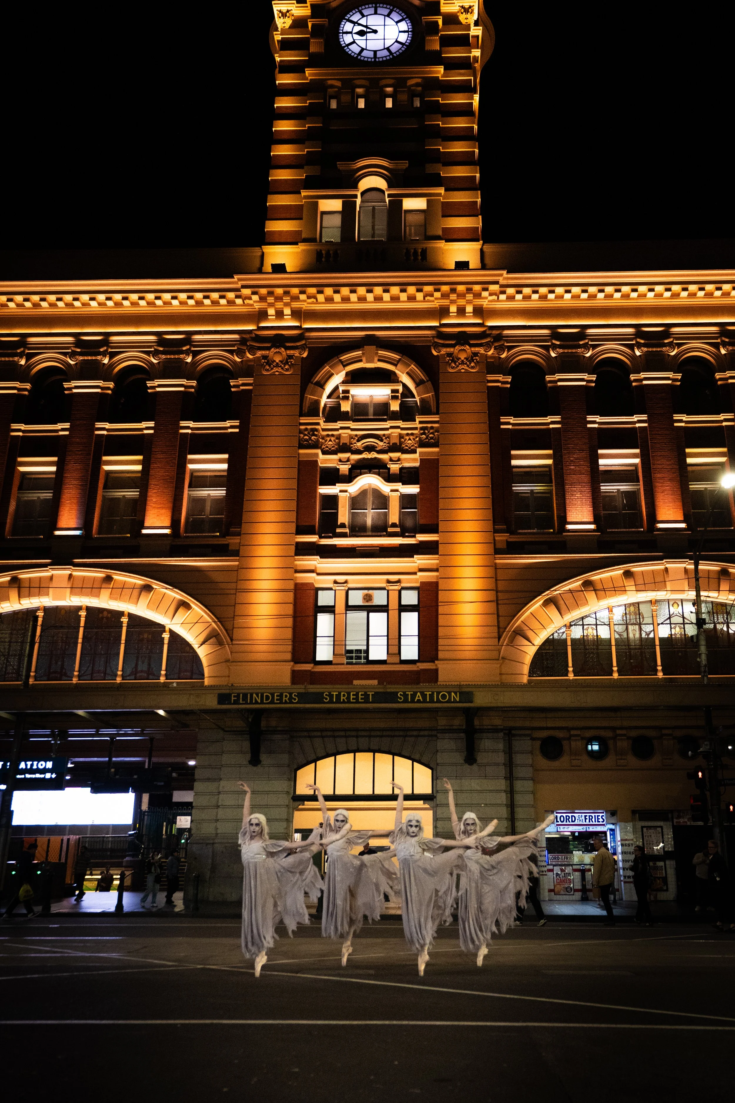 Night view of Flinders Street Station in Melbourne illuminated with warm lights, with four dancers dressed as ghosts in white costumes performing in front of the station.