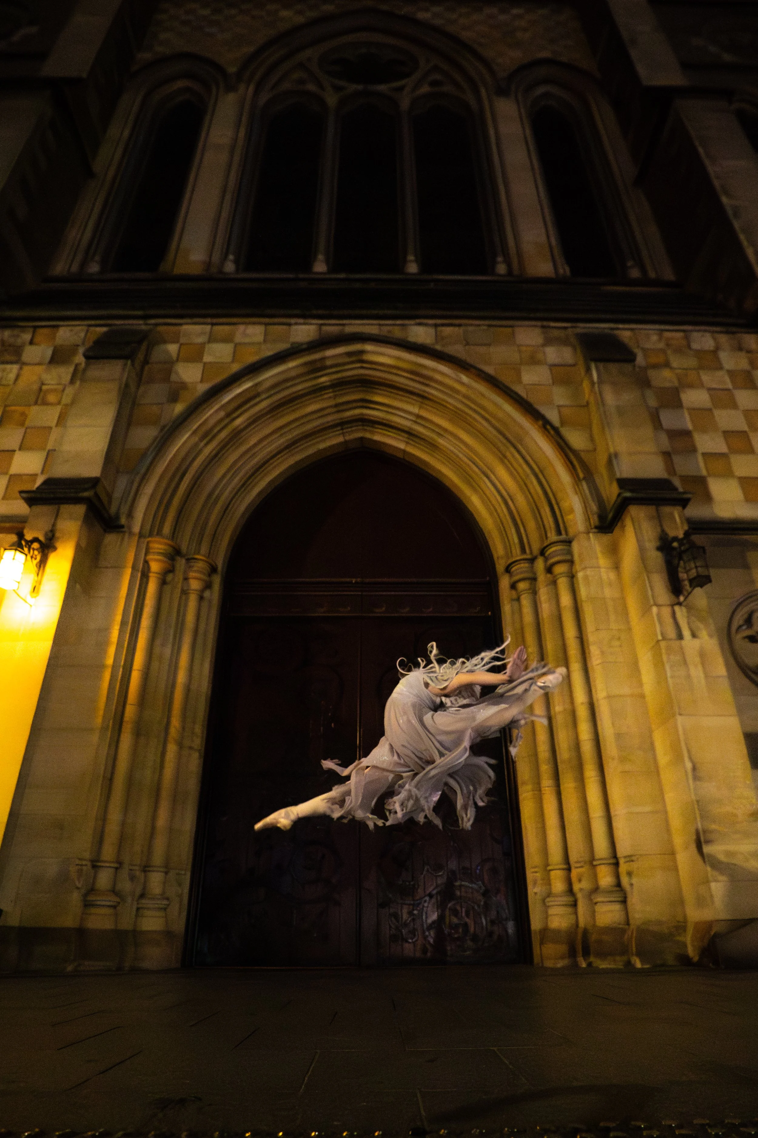 Nighttime view of a Gothic-style church with tall stained glass windows and an ornate stone facade. A white angel sculpture with flowing robes and wings is positioned in front of the entrance, illuminated by exterior lights.
